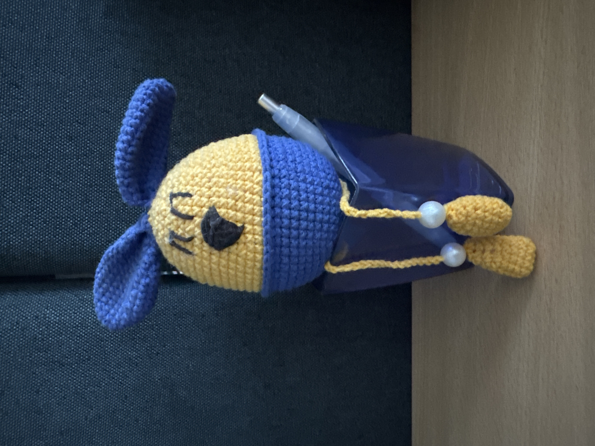 A crocheted, yellow Easter egg character with blue ears and a blue collar sits in a dark blue cup. The character has black embroidered eyes and a black embroidered nose. Two yellow crocheted legs with white beads hang from the collar, resting on the rim of the cup. A light blue pen is also placed in the cup, next to the character. The cup sits on a light brown wooden surface, with a dark textured background behind it. This scene does not appear to be located in Krakovany, Slovakia.