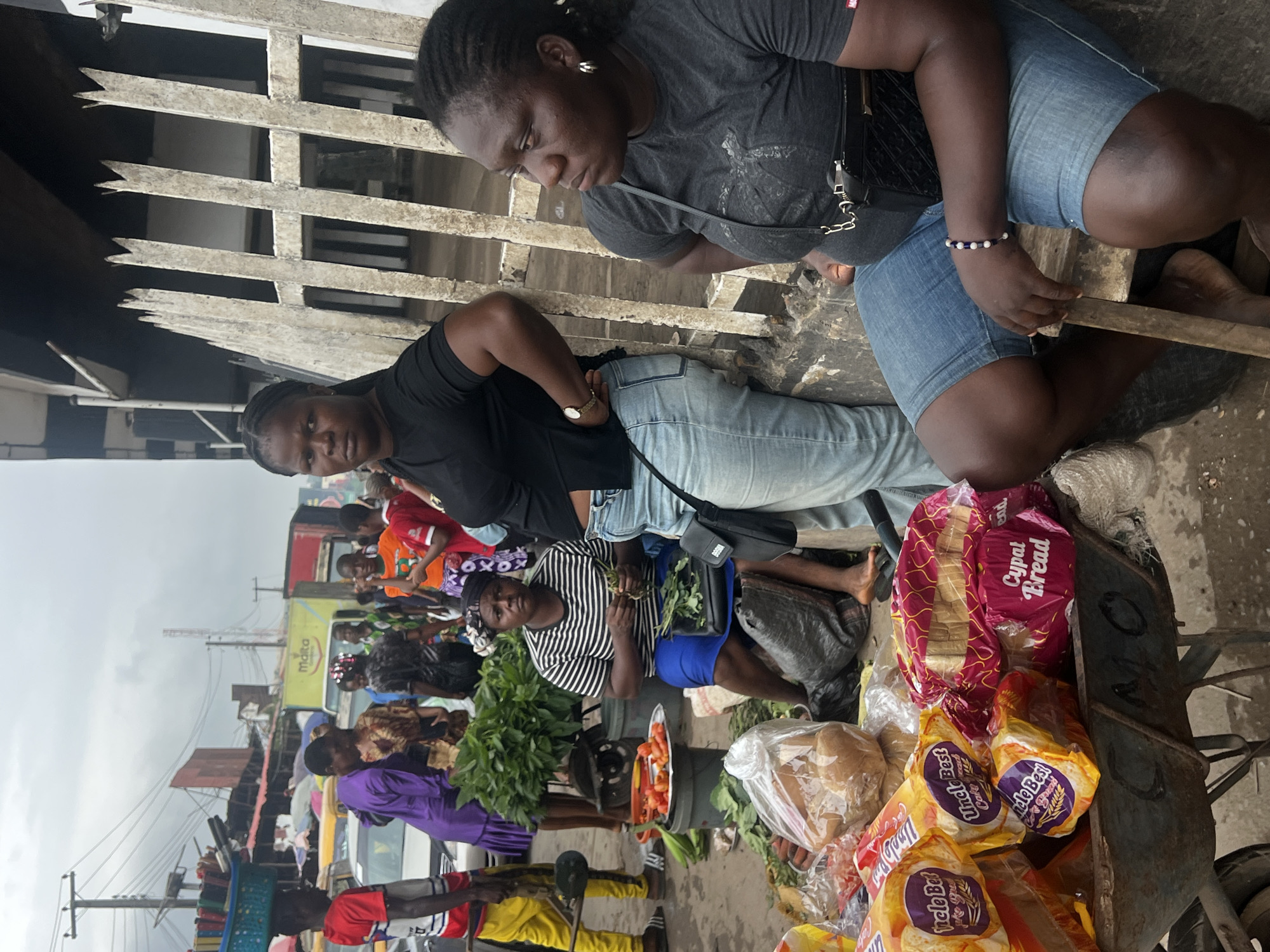 The image depicts a street scene in Nkpor, Nigeria, likely a marketplace given the presence of vendors and a variety of goods. In the foreground, a person is seated behind a wheelbarrow filled with bread loaves and bagged produce. To their left, another individual is sitting, preparing vegetables. Further back, several other people are visible, some appearing to be vendors or customers. They are dressed in casual attire. The environment shows a paved street, with some buildings and market stalls in the background, along with vehicles. The sky is overcast, suggesting a cloudy day. The overall impression is one of a bustling urban commercial area.