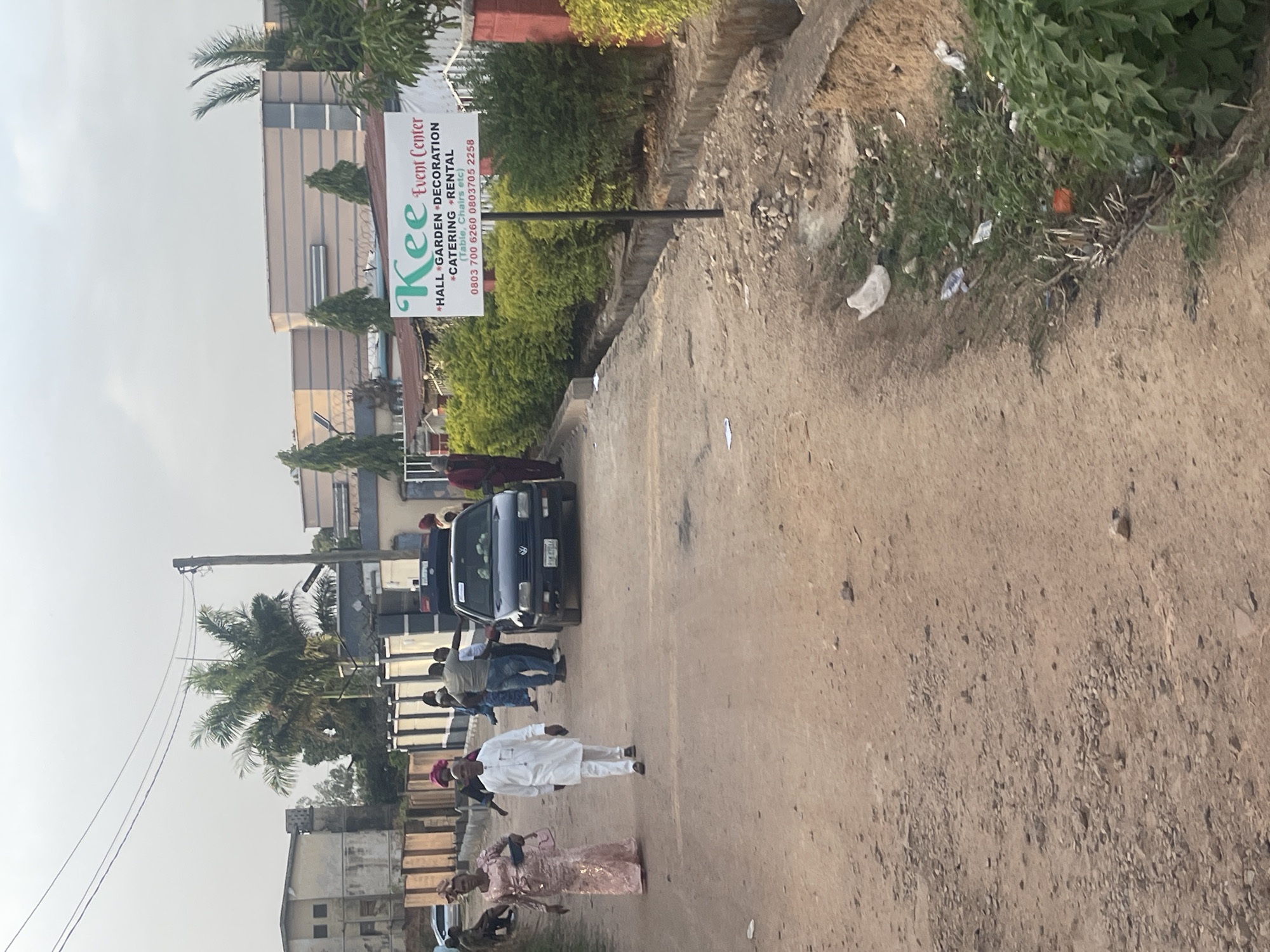 The image depicts a street scene in what appears to be Jos, Nigeria, indicated by the phone numbers on the "Kee Event Center" sign. The dirt road is flanked by vegetation and buildings. A group of people is walking along the road, and a dark-colored vehicle is parked nearby. The event center sign advertises hall/garden decoration, catering, and rental services. The overall scene suggests a casual, outdoor event or gathering.