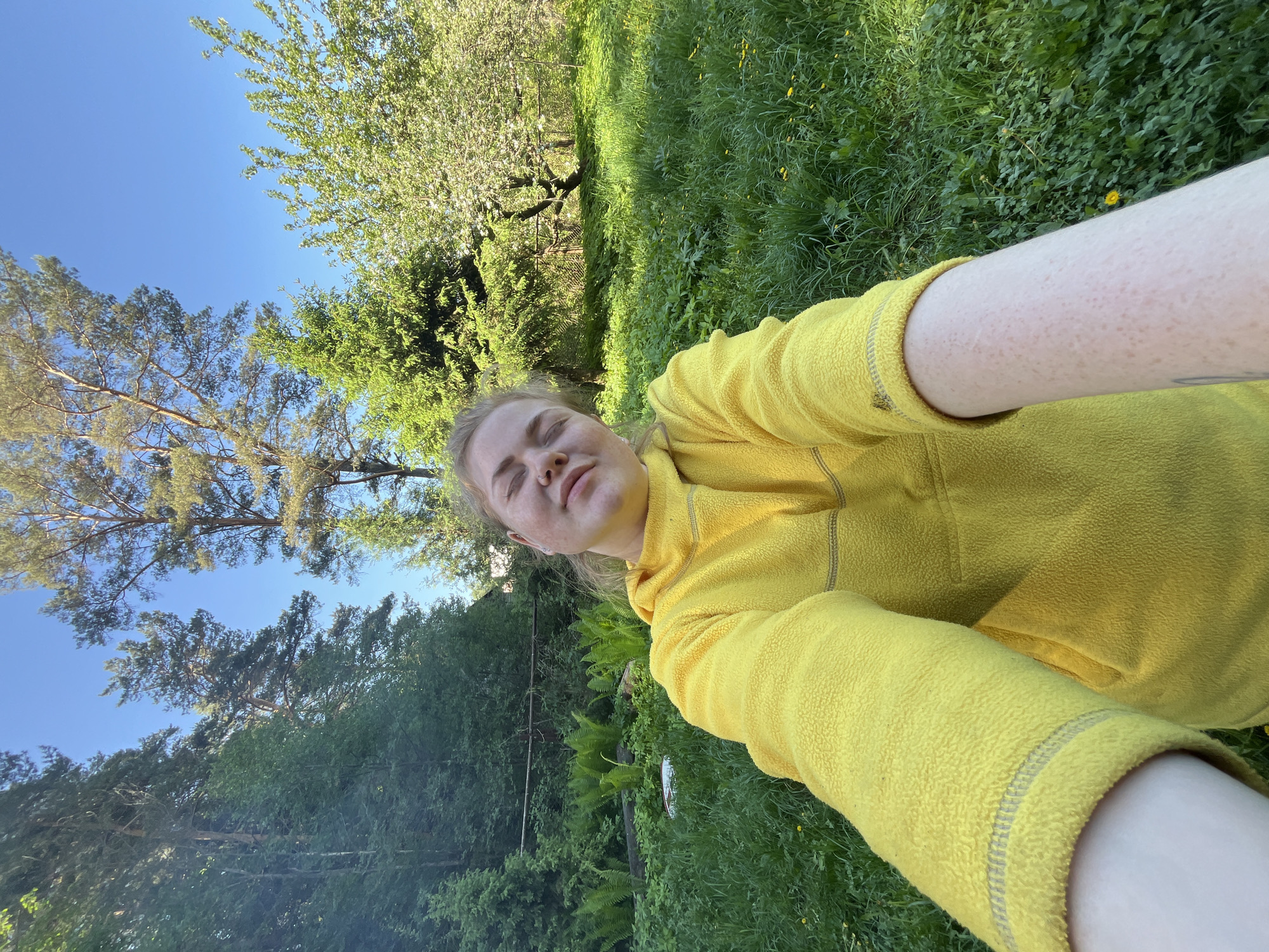 A young woman wearing a yellow fleece jacket is lying on her back in a grassy area in Chaikovsky, Russia. She has her eyes closed and appears to be relaxed or sleeping. The sky is blue and visible through the trees, which are a mix of deciduous and evergreen varieties. There are ferns growing in the grass. The perspective of the photo suggests it was taken as a selfie.