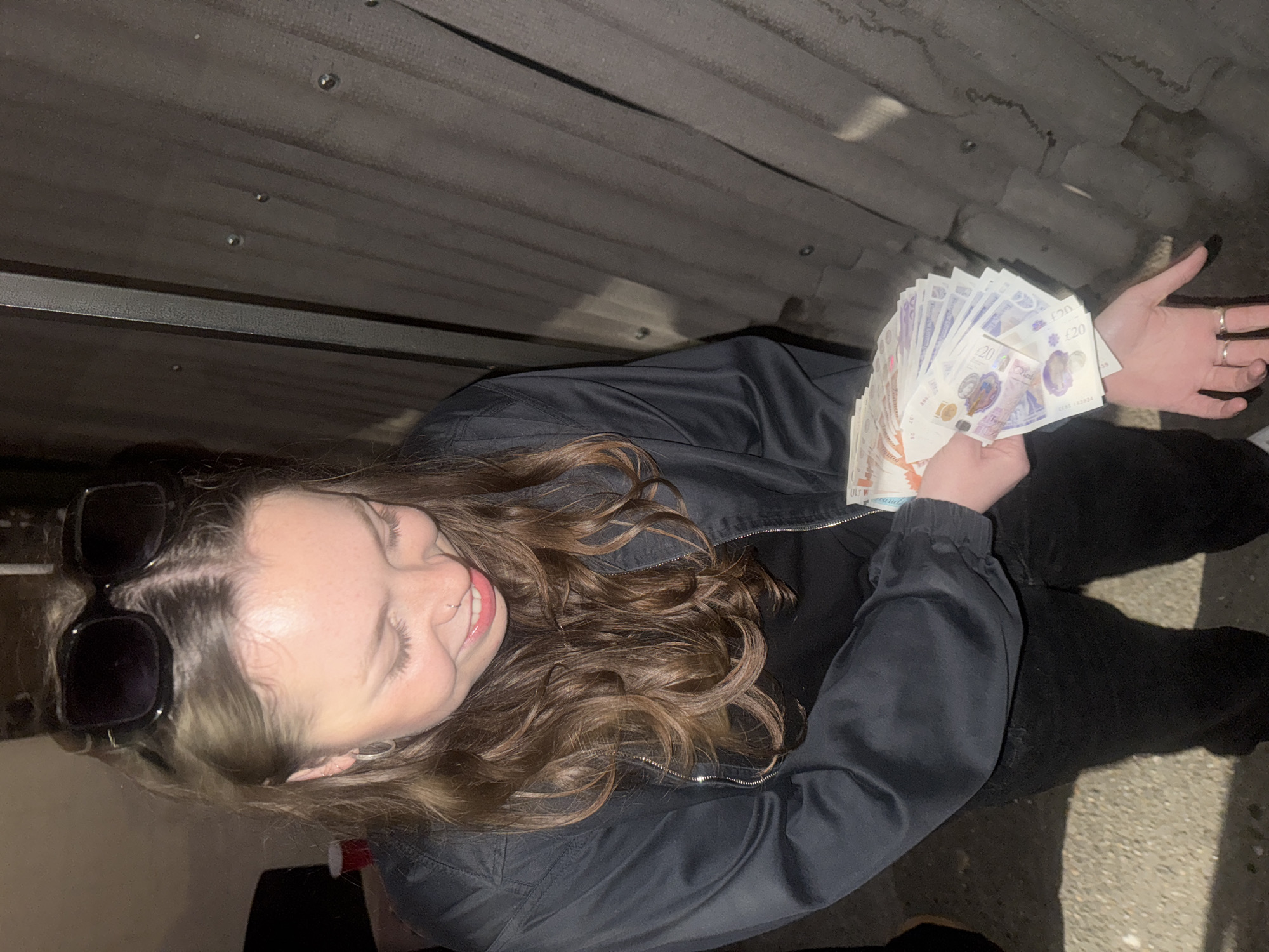The image, taken from a high angle, shows a young woman posing with a fanned stack of British twenty-pound notes. She has long, wavy brown hair and a light complexion, captured mid-smile, conveying a cheerful or satisfied expression. She is wearing a dark, possibly black, zipped jacket over a dark top, with large dark sunglasses pushed up onto her head. Her left hand holds the cash, clearly displaying the "£20" denomination, the image of Queen Elizabeth II, and the "BANK OF ENGLAND" text on the polymer banknotes. Her right hand, open, reveals two rings on her fingers.
The setting appears to be an urban or industrial environment in London, United Kingdom. The background is a dark, corrugated metal surface, likely a wall or ceiling, suggesting an underpass, an alleyway, or a covered area with limited light. The ground visible at the bottom right is dark, possibly concrete or asphalt. The overall lighting is dim, indicating it might be evening, night, or an enclosed space. The woman's pose and broad smile suggest she is deliberately showcasing the money for the photograph, potentially celebrating a financial gain or simply posing with cash.