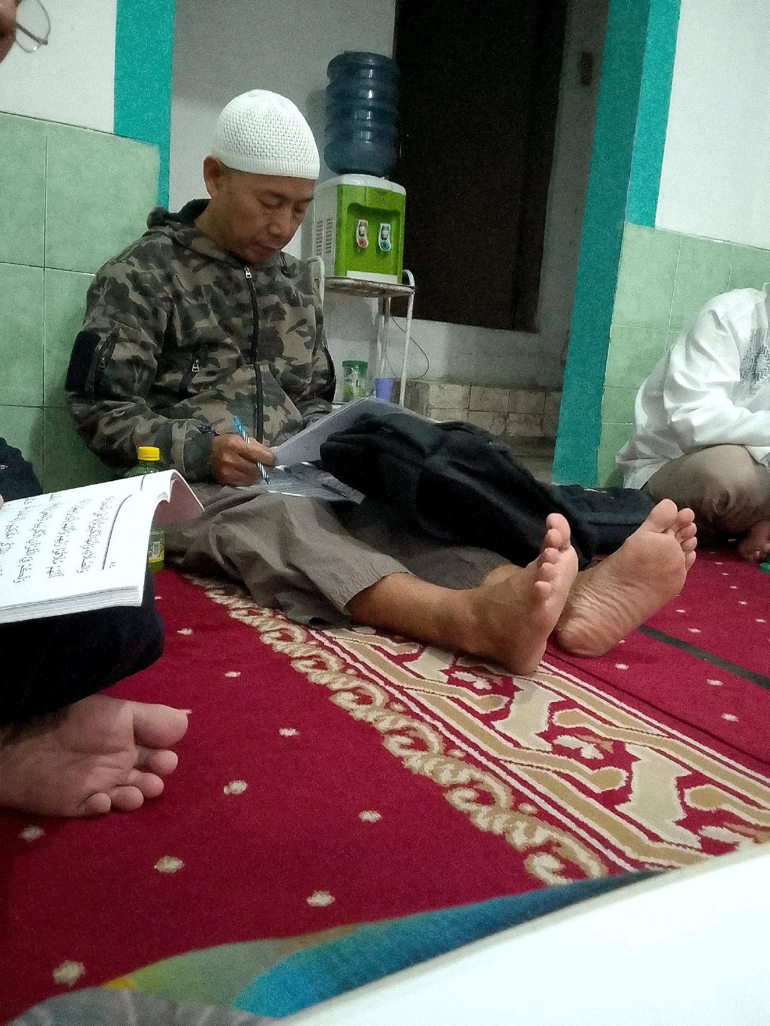 In Gunungmasigit, Indonesia, a man wearing a white skullcap and a camouflage jacket sits on a red carpet with intricate beige and white patterns. He is focused on a book open before him, writing in it with a blue pen. His legs are crossed, and his feet are visible. Another individual in a white shirt is partially visible in the background. The environment appears to be indoors, with tiled walls in shades of green and white, and a water dispenser visible behind the seated man. The lighting is dim, suggesting it might be evening or an interior space with limited natural light.