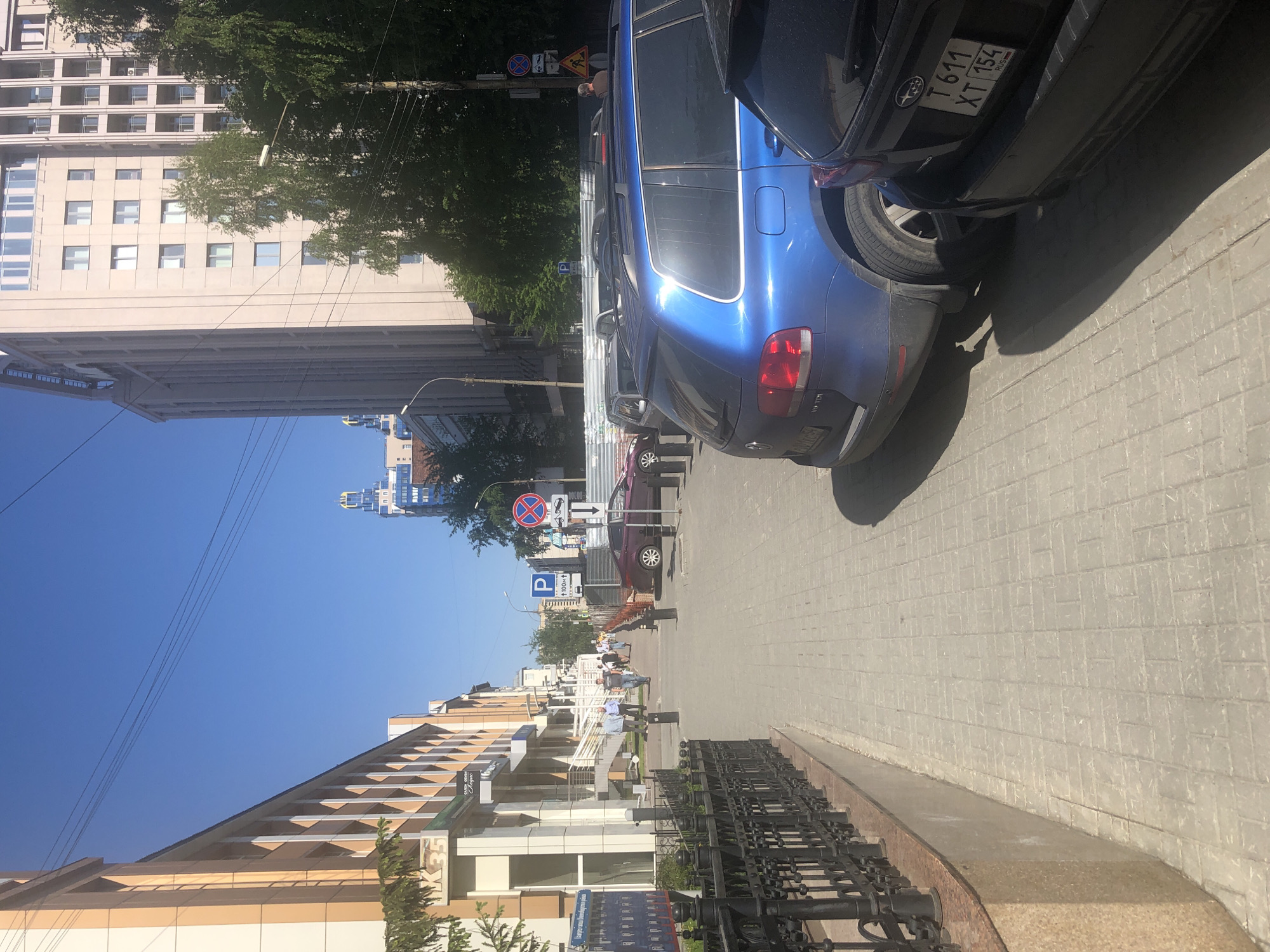 The image depicts a street scene in an urban area. On the right side of the frame are parked cars, including a blue SUV and a dark-colored car with the license plate "T 611 XT 154." The street is paved with rectangular blocks. On the left, there are buildings with varying architectural styles, including a tall beige building with numerous windows. Street signs are visible, including a "No Stopping" sign and a "Parking" sign. Further down the street, there are people walking. The sky is clear and blue. A black metal fence is situated along the lower left of the image.