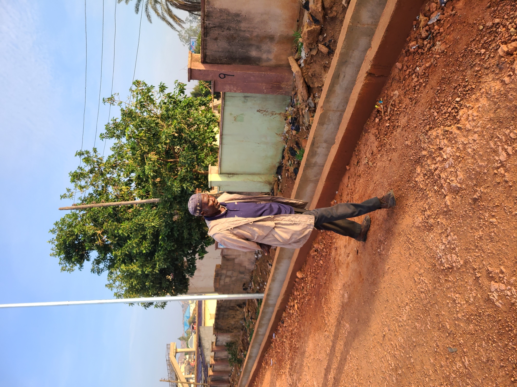 An elderly man stands on a dirt road in Jos, Nigeria. He wears a blue shirt, dark trousers, a light tan jacket, and a dark patterned cap. The road is reddish-brown dirt with scattered rocks. A concrete curb runs alongside the road. Behind the man, there's a green tree and several buildings, including one with a light green facade. In the distance, there's infrastructure, possibly a bridge or elevated road. Power lines run across the top left of the frame against a blue sky. There is garbage visible between the curb and the buildings. The light indicates it may be early morning or late afternoon.