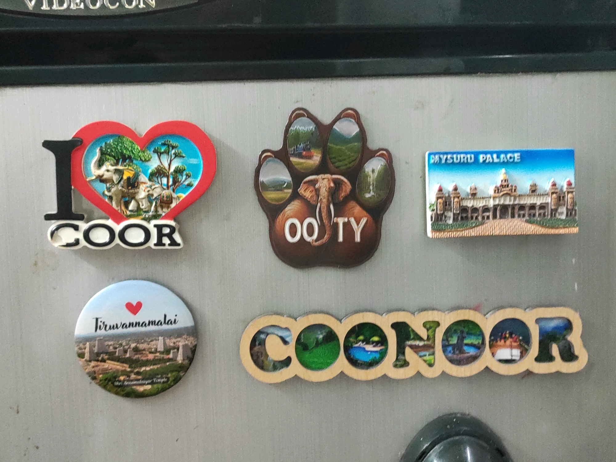 The image displays a collection of magnets on a refrigerator door. The magnets feature various tourist destinations. From left to right, top to bottom: a magnet with an elephant and trees inside a heart shape, labeled "I COOR". Next to it is a paw print magnet with scenic views and "OO TY" written at the bottom. To the right of that is a magnet depicting Mysore Palace with the text "MYSURU PALACE". Below the "I COOR" magnet is a circular magnet with a heart and the text "Tiruvannamalai", showing a cityscape. Finally, a wooden cutout magnet spells out "COONOOR" with small scenic pictures inside each letter. The refrigerator door is a metallic gray, and the top of the image shows the edge of a dark appliance. The scene is indoors, likely in a kitchen. There are no people or animals present in the image. The overall presentation suggests personal mementos from travels. This collection of magnets suggests the refrigerator is located in a residence, as it's common for people to decorate their refrigerators with such items. While magnets can indicate personal interest in specific locations, without further context from the arrangement or additional items in the environment, it's impossible to definitively determine the exact location within Hyderabad, India, or any other city.