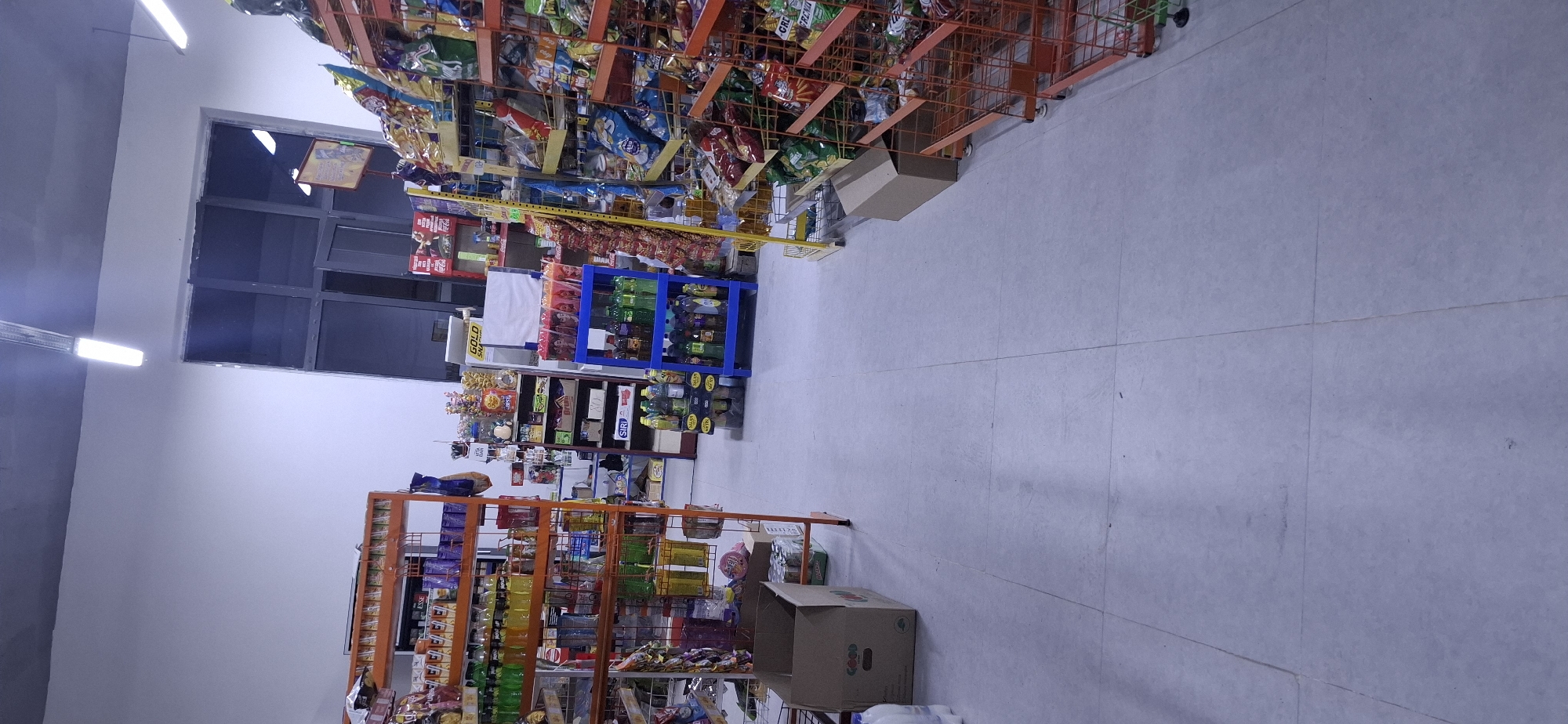 The image shows the interior of a small convenience store located in Arys, Kazakhstan. The store is stocked with various goods, including snacks, beverages, and other grocery items displayed on metal shelves. The floor is tiled with light-colored squares. In the foreground, a cardboard box and some cans are visible. In the background, there is a large window or door, and fluorescent lighting fixtures are mounted on the ceiling. The store appears to be well-lit and organized. There are no people visible in the image.