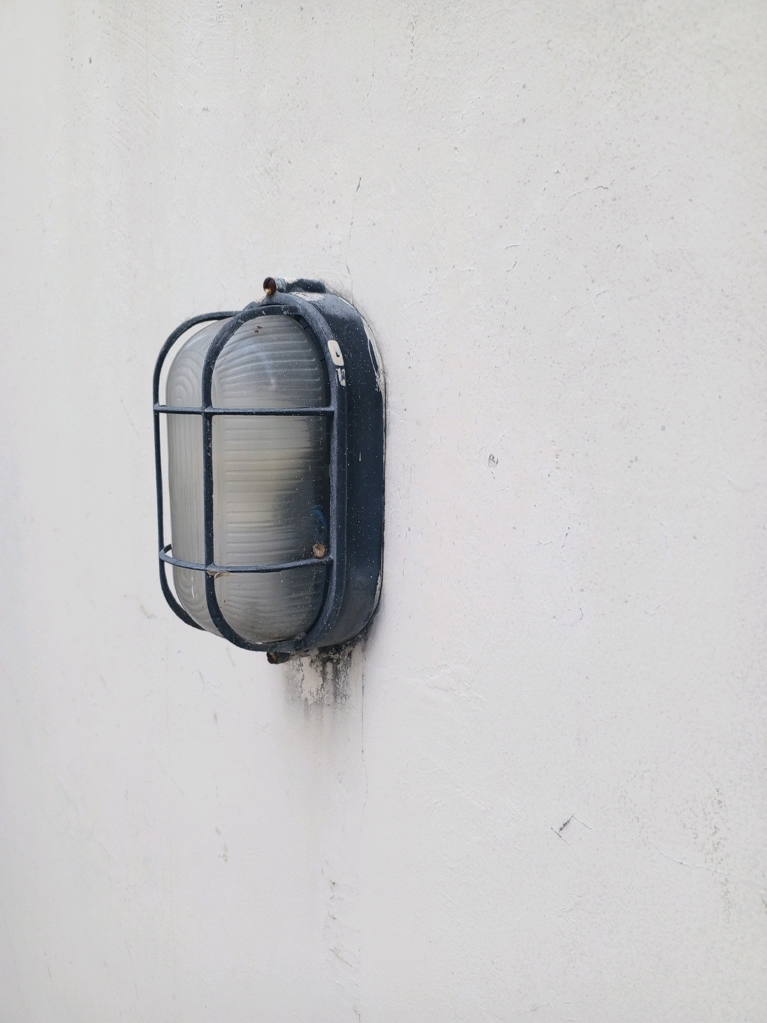 An outdoor bulkhead light fixture is mounted on a rough, white-painted concrete wall. The light fixture is dark gray with a protective metal cage over a ribbed glass lens. The wall shows some signs of wear, with minor cracks and discoloration, particularly below the light fixture, suggesting it is exposed to the elements. The scene is devoid of any human presence or activity. The lighting is neutral, indicating it is daytime. The image composition focuses on the texture of the wall and the details of the utilitarian light fixture. It is impossible to determine the precise location within Denpasar, Indonesia, based solely on this image.