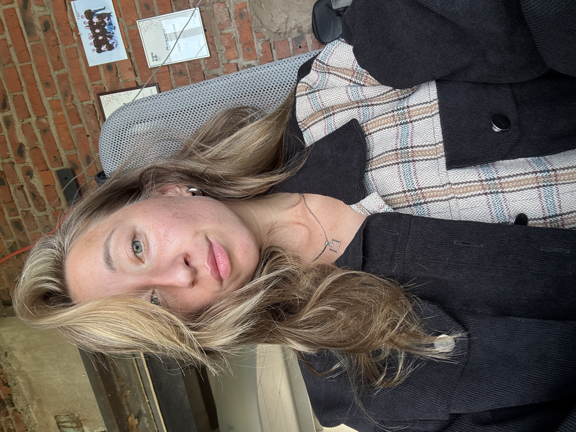 A young woman with blonde hair pulled back slightly and framing her face, is captured in a close-up selfie taken from a slightly low angle. She is seated, wearing a split jacket: one side is black corduroy with a button closure, and the other features a plaid pattern with shades of beige, white, and subtle blue and red lines. She wears a simple silver necklace with a rectangular pendant. Her gaze is directed towards the camera, her lips slightly parted. The background consists of a brick wall, with a wire extending from the top left corner downwards, and a metal-framed office chair visible to her right. Several framed items are mounted on the brick wall, including what appear to be certificates and a group photograph. The lighting suggests an indoor setting, possibly an office or studio, with a window or light source to the left. The overall impression is a candid shot.