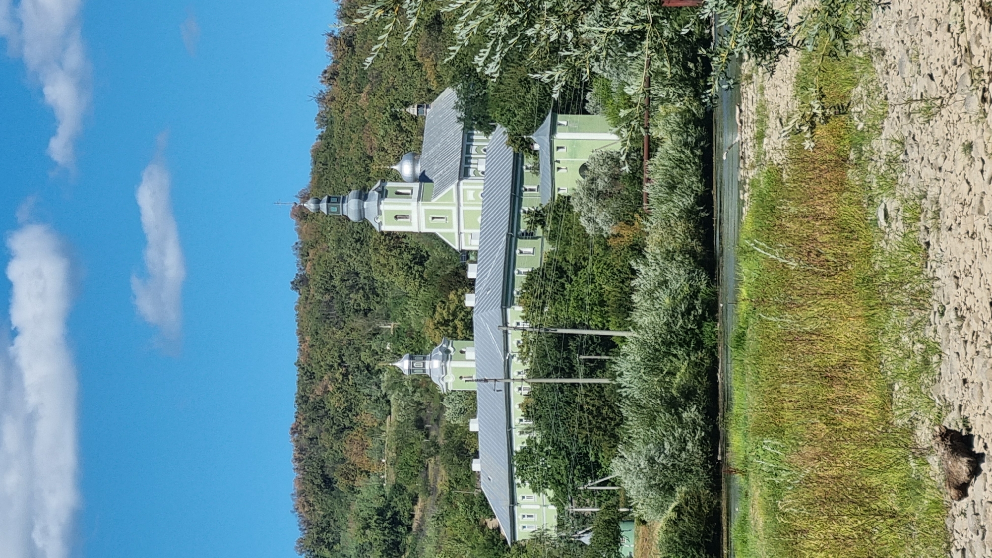 The image shows a large, light green monastery complex nestled amongst rolling, tree-covered hills under a clear blue sky with scattered white clouds. The monastery features multiple buildings with gray, sloped roofs, several prominent domes, and at least two bell towers. One tower is taller and more elaborately designed, topped with a spire and cross. The complex appears to be situated on a slight incline, with the buildings directly facing the viewer. In the foreground, a body of water, likely a river, is visible, bordered by a bank with tall, dry grass and rocks. Dense green foliage and trees surround the monastery and the riverbank, creating a natural, slightly wild landscape. A few utility poles with wires are visible in front of the monastery buildings. The overall impression is one of a serene, historic religious site in a verdant, natural setting.
