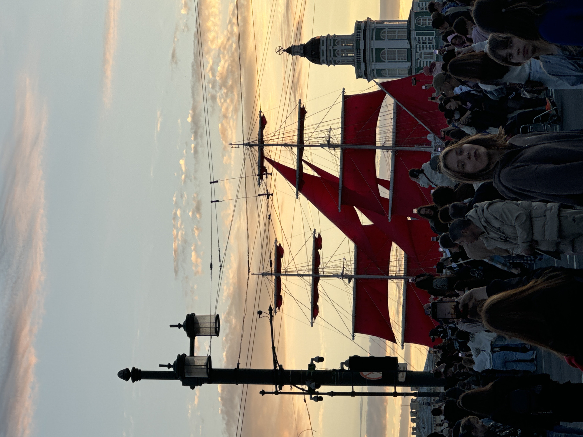 In Saint Petersburg, Russia, a crowd of people gathers along the Neva River during the "Scarlet Sails" festival. The iconic tall ship, the "Shtandart," with its striking red sails, is prominently featured in the background. The evening sky is painted with warm hues of pink and orange, casting a soft glow over the scene. People are seen holding up their phones to capture the spectacle, their faces illuminated by the device screens. A lamppost with multiple lights stands to the left, and a historical building with a dome-shaped tower is visible behind the ship. The atmosphere is festive and celebratory, with the magnificent ship and the vibrant sky creating a memorable backdrop for the onlookers.