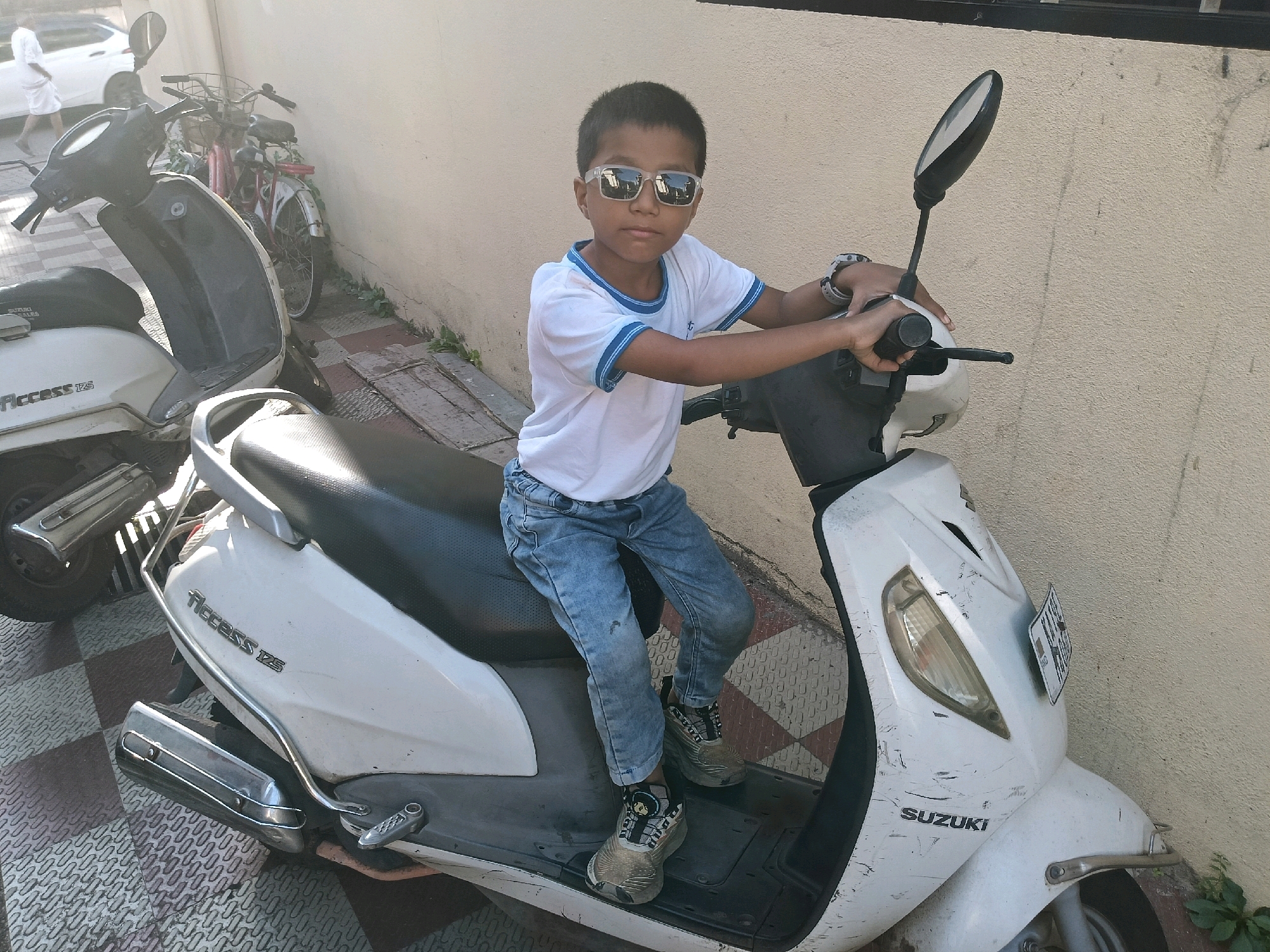A young boy wearing sunglasses, a white t-shirt with blue trim, and blue jeans sits on a white Suzuki Access 125 scooter. He is holding the handlebars and looking towards the camera. The scooter is parked next to a textured, light-colored wall. In the background, other parked vehicles including a bicycle and another scooter are visible. The ground is paved with interlocking tiles. The scene appears to be outdoors, possibly in a residential or commercial area in Mangaluru, India, given the scooter model and common urban setting.