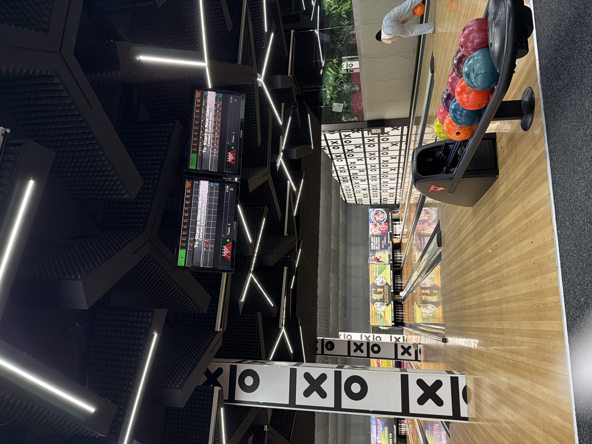 The image shows a bowling alley, possibly in Burgas, Bulgaria based on the Cyrillic advertisements. It features bowling lanes, bowling balls in a rack, a person in the background preparing to bowl, and monitors displaying scores. The decor includes black walls with geometric acoustic panels, linear lighting fixtures, and decorative elements featuring "X" and "O" patterns. There are advertisements on the walls along the lanes.