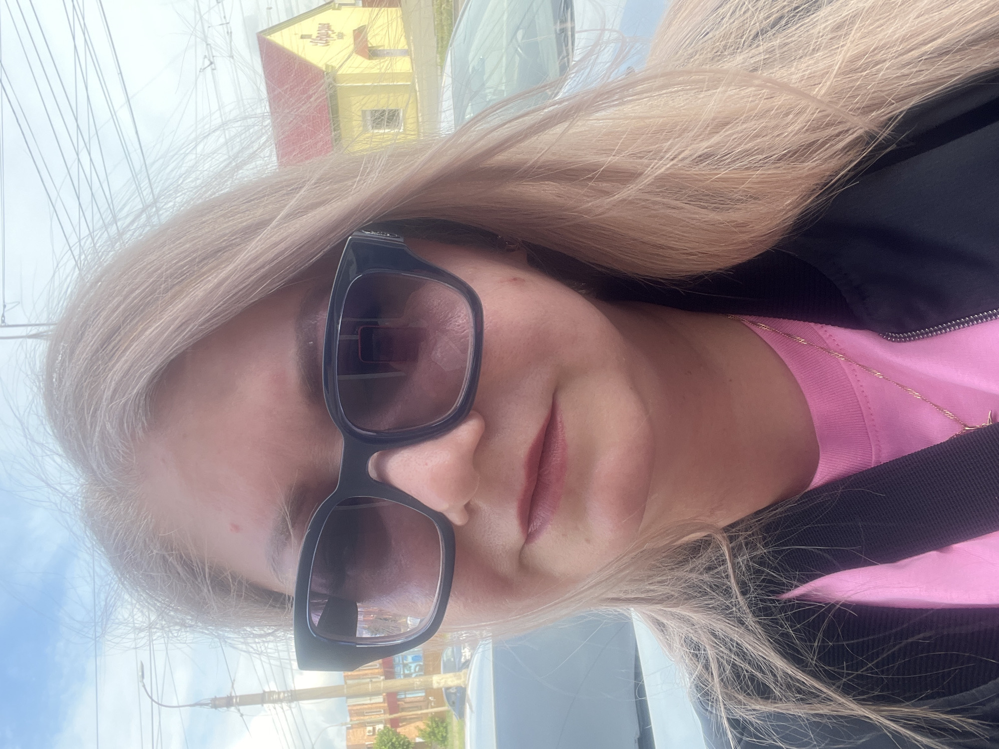The image is a selfie of a woman with long, straight, light blonde hair. She is looking directly at the camera, wearing large, black-framed sunglasses that cover her eyes. She has a subtle pink lipstick on her lips. She is dressed in a bright pink top, with a black jacket or coat partially visible over it, and a delicate gold chain is worn around her neck.
The setting is outdoors, likely an urban or suburban street in Izhevsk, Russia. In the background, particularly visible in the reflection on her sunglasses, there is a yellow building with a red roof. On the facade of this building, Cyrillic text is discernable, which reads "ПРОДУКТЫ" (Russian for "Groceries" or "Products"), indicating a commercial establishment. Overhead, numerous power lines or utility wires are visible against a bright, hazy sky. Parts of other vehicles are also reflected in the sunglasses and can be faintly seen in the background. The bright, even lighting suggests it is daytime. No specific activity beyond taking the selfie is depicted, and her emotions are not discernible due to the sunglasses.