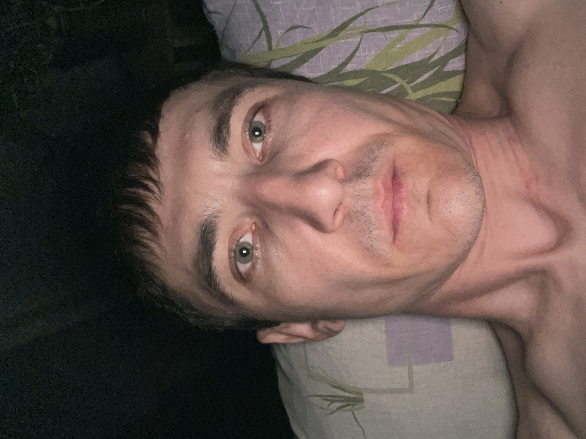 A close-up, eye-level selfie of a man's face is captured in dim lighting. The man is lying down, as indicated by his head resting on a pillow. His body is partially visible from the shoulders up. He has short, dark hair with bangs that fall across his forehead. His eyes are open and looking directly at the camera, with green irises. He has a fair complexion with some redness on his cheeks and around his nose. His lips are slightly parted, revealing a hint of his teeth. A light stubble is visible on his chin and jawline. The pillow beneath him has a light-colored floral pattern with green leaves and purple squares. The background is dark and out of focus, suggesting an indoor setting at night. There are no other people or objects of significance in the frame. The image does not provide any specific details about the city of Laishevo, Russia.