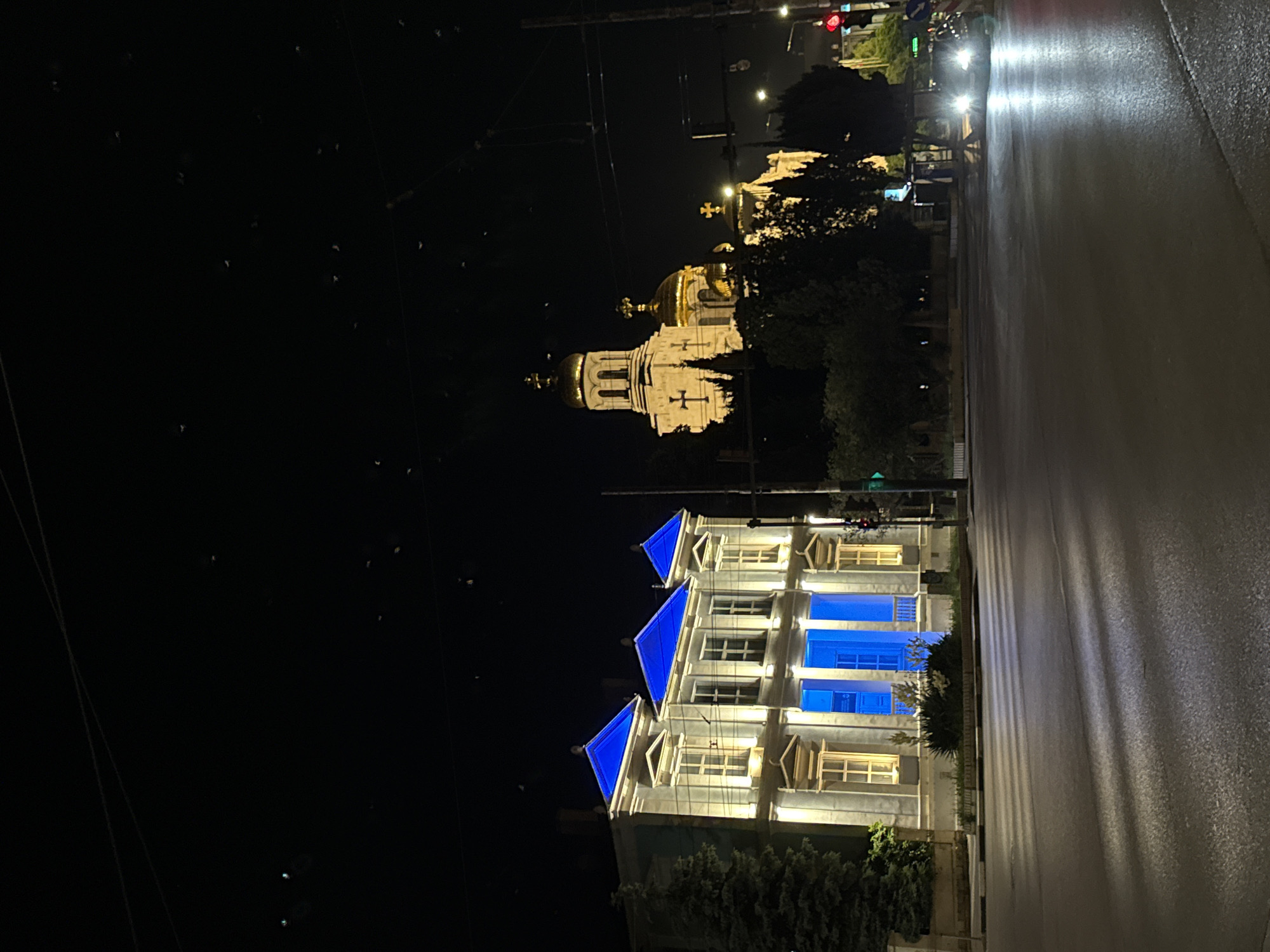 This image depicts a nighttime scene in the city of Varna, Bulgaria. On the left, a two-story building with a classical facade is illuminated by yellow lights, with its roof and central section bathed in bright blue light. This building appears to be an official or cultural institution. To the right and slightly behind it, the golden domes of a large, ornate church rise against the dark sky. The church features intricate architectural details and crosses atop its domes.
The foreground shows a wet asphalt road reflecting the lights from the surrounding buildings and streetlights. A single car with its headlights on is visible turning at an intersection, its light beams creating long reflections on the wet surface. Traffic signals are present, with a red light visible on the right side of the frame. The overall atmosphere is one of urban nighttime ambiance, with the illuminated buildings serving as prominent landmarks. The specific locations of the buildings are identifiable as the Varna City Hall (on the left) and the Dormition of the Mother of God Cathedral (on the right).