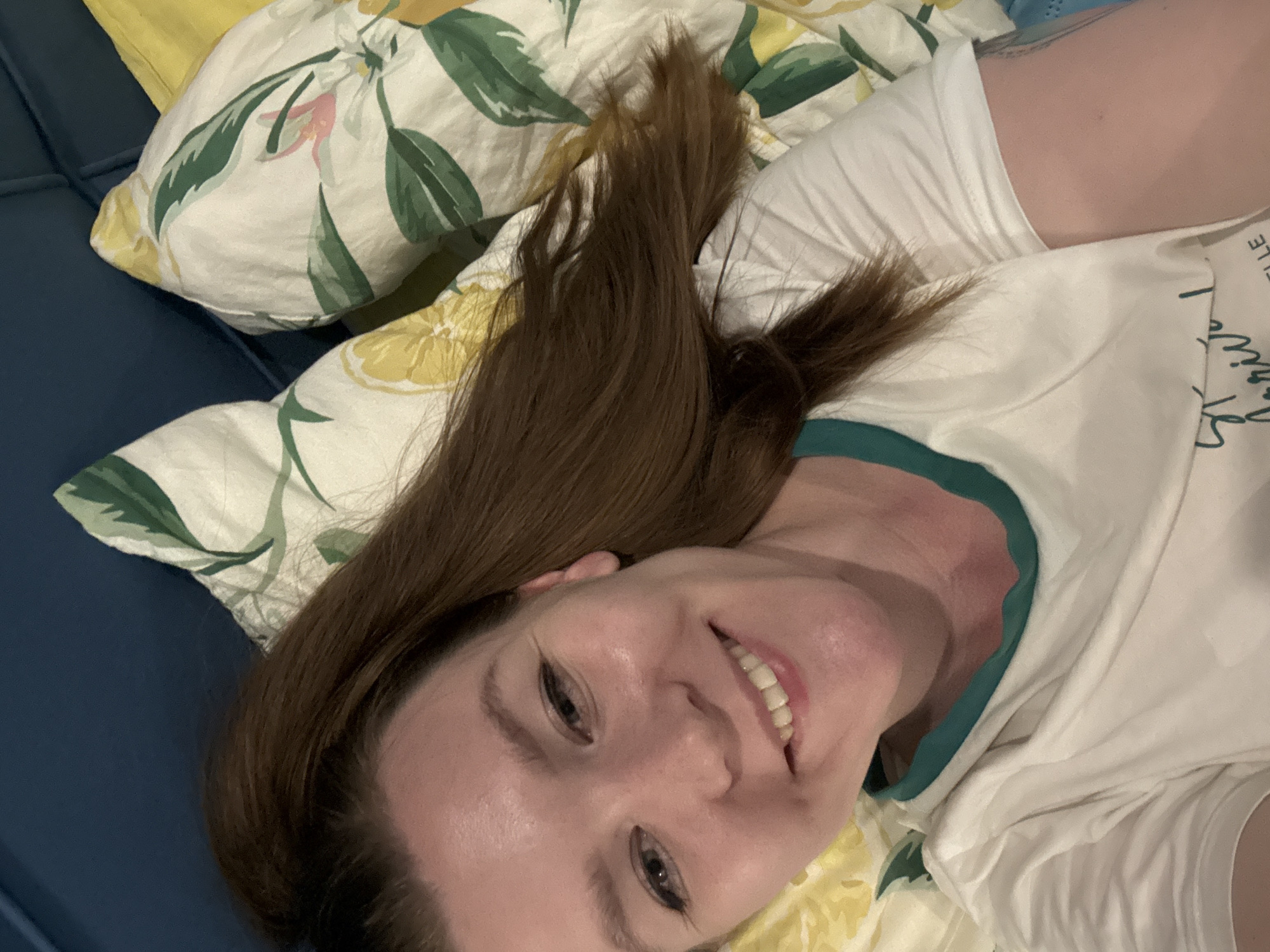 A woman is lying on a bed. She has medium-brown hair and is smiling. She is wearing a white t-shirt with a green collar. The bed has a blue headboard and yellow and white floral patterned pillows. There is not enough information to determine if the image was taken in Izhevsk, Russia.