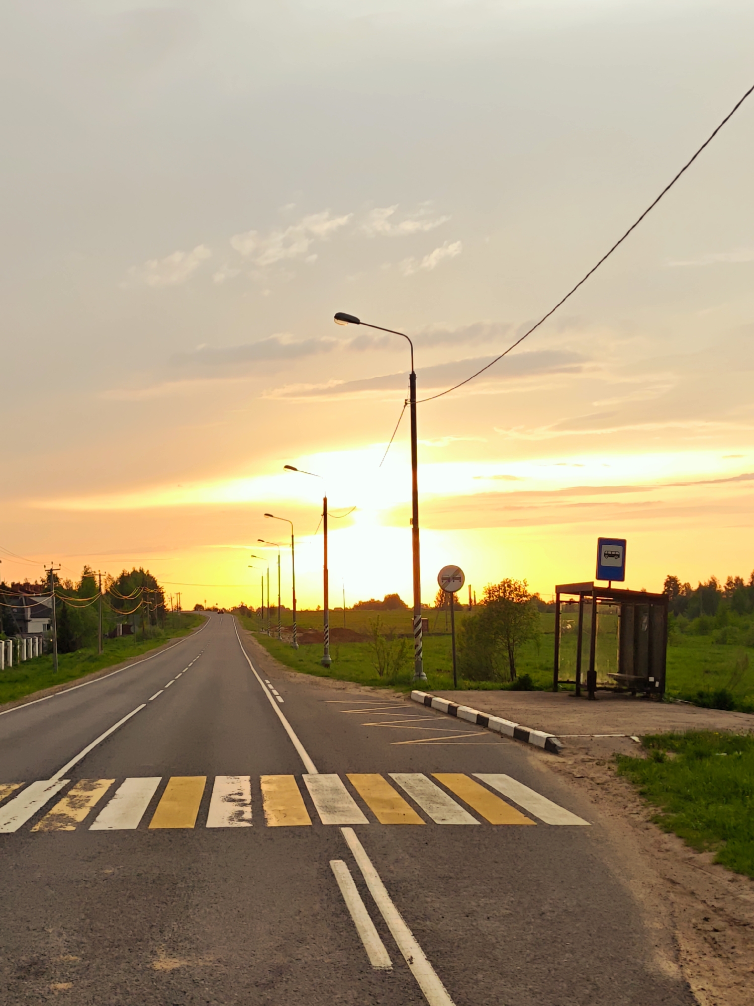 The image shows a view of a road in Курово, Russia at sunset. The road stretches into the distance, lined with streetlights. A crosswalk with alternating yellow and white stripes is visible in the foreground. To the right of the road is a bus stop with a bus symbol on top. Vegetation is visible on both sides of the road. Overhead electrical wires span across the sky. The sky is filled with colors of orange and yellow from the setting sun.