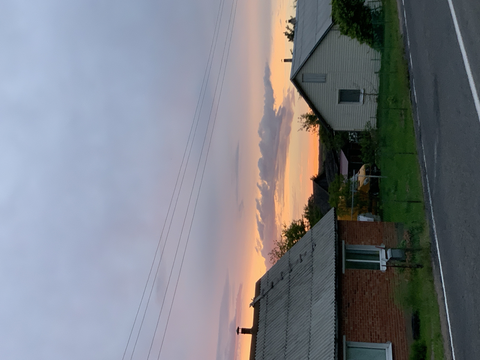 The image captures a rural scene at dusk, with a colorful sky featuring hues of orange, pink, and purple. Several houses are visible, including a brick house with a corrugated metal roof on the left and a lighter-colored house with a similar roof on the right. Power lines stretch across the sky. The foreground shows a road with a white line marking its edge. The environment appears to be a village or small settlement, potentially in the area of Відзаўскі сельскі Савет, Belarus, given the rural architecture and landscape. No people are present in the image.