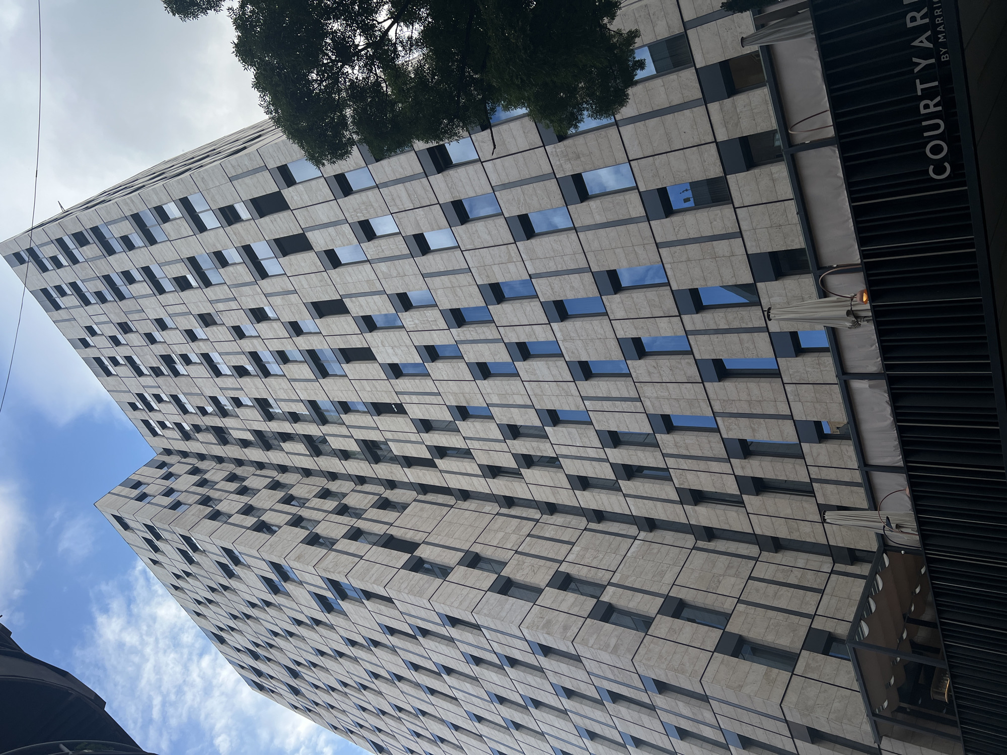 The image shows a tall building in the city of Unknown, Unknown. The building has a distinctive facade with a pattern of rectangular windows and stone or concrete panels. A sign with the words "COURTYARD BY MARRIOTT" is partially visible on the right side of the building. A tree is partially visible at the top of the building. The sky is visible with blue and white colors.