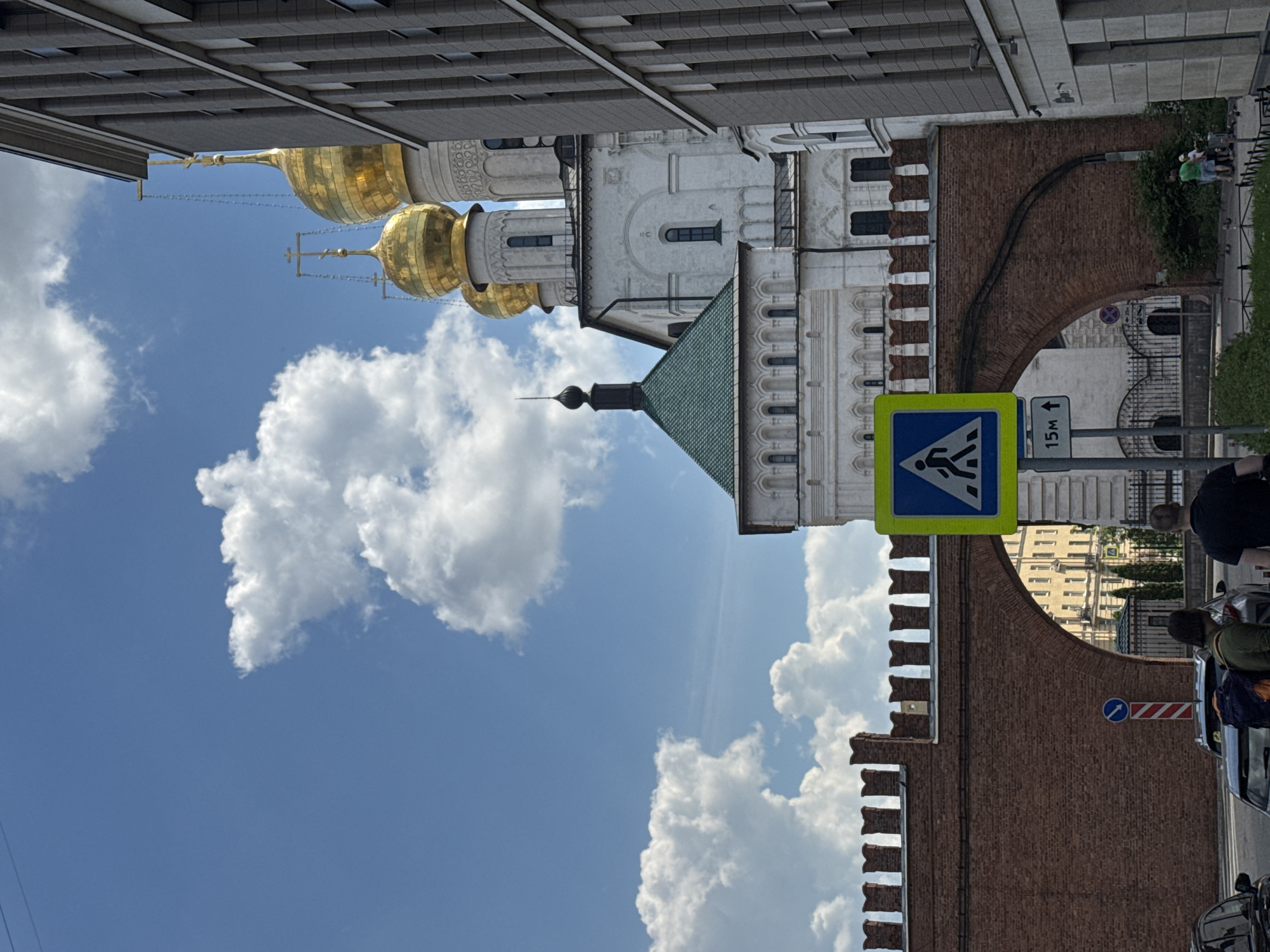 The image presents a daytime urban scene under a bright blue sky adorned with fluffy white clouds. The central focus is on a collection of diverse architectural styles. In the mid-ground, two prominent white or light-colored buildings, possibly churches or cathedrals, showcase traditional architecture with multiple golden onion domes, each topped with a cross. Another building with a dark green, sloped roof and a slender spire is also visible among these structures. Contrasting with these historical elements, a modern building with a dark, gridded facade spans the upper portion of the frame.
A significant reddish-brown brick wall, featuring a distinctive crenellated top and an arched opening, diagonally crosses the lower and mid-sections of the image, partially obscuring background details. At street level in the lower right, a few people are visible; one person wearing a green shirt stands near green bushes, and another with dark hair is next to a silver car. Part of a black car is also seen.
Several road signs are prominent in the foreground: a bright yellow and blue pedestrian crossing sign with a walking figure, and below it, a white rectangular sign displaying "15m" with an upward arrow. Further back, near the brick wall, a blue circular sign with a white arrow pointing right and a red and white striped barrier are visible. Thin wires or cables crisscross the sky near the golden domes. The scene suggests a sunny day in a historical urban environment, with the distinctive onion domes strongly indicating a Russian context.
