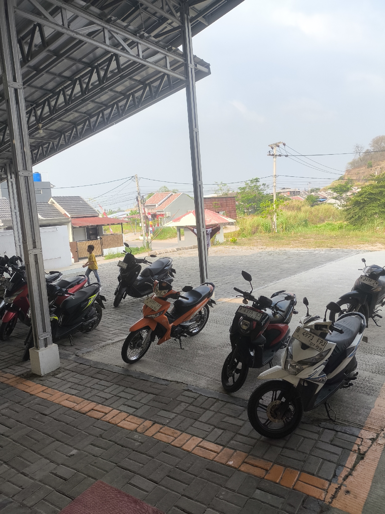 In Cilegon, Indonesia, under a covered walkway with a metal truss roof, multiple parked motorcycles are visible. The ground is paved with interlocking bricks, featuring an orange stripe. A young boy in a yellow shirt walks away from the camera towards a building with a red-tiled roof in the background. Beyond the building, a wooded hillside with some houses and utility poles is apparent under a hazy sky. Several of the motorcycles have license plates.