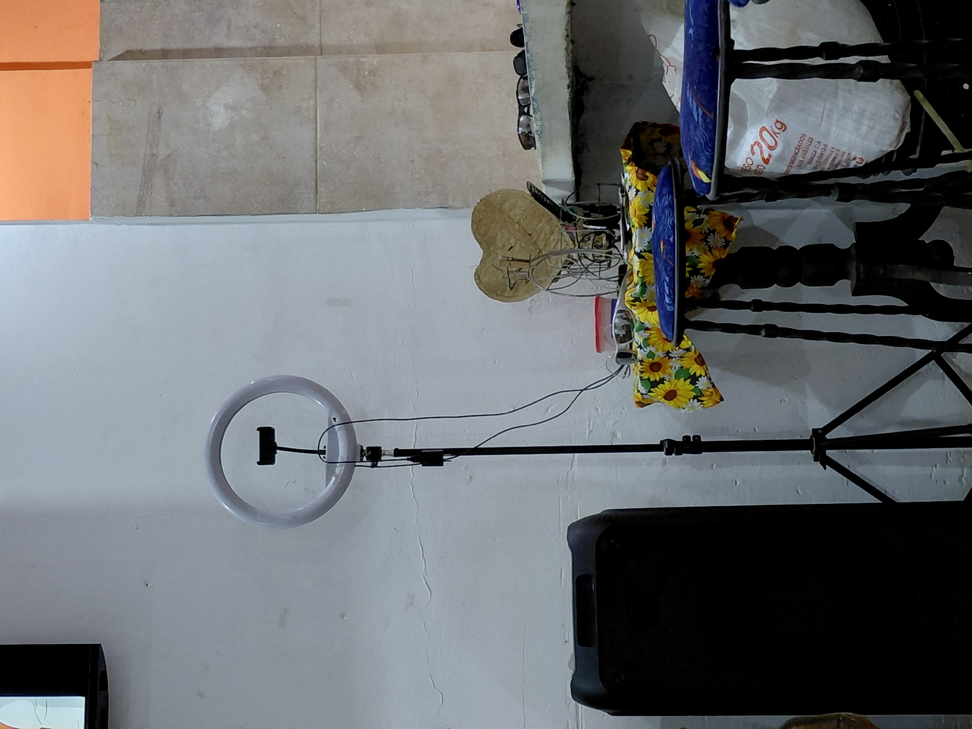 The image shows a room interior with a ring light on a stand in the lower left. A black speaker is positioned to the right of the ring light. Above these items, against the wall, is a hat, a table with a sunflower-patterned cloth draped over it, and two blue chairs. A bag of flour (20kg) is visible near the chairs. The wall is painted white. The top of the image shows a tiled floor. The location is Santa Ana, Venezuela.