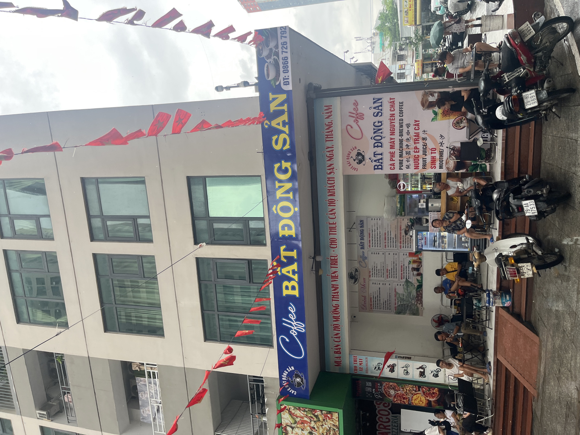 The image captures a vibrant street scene at a coffee shop and real estate business in Bac Nha Trang Ward, Vietnam, during the daytime, likely under an overcast sky. Several individuals, mostly men, are seated at small tables on the sidewalk, enjoying drinks and socializing. Most are casually dressed, some using their phones. Motorcycles and scooters are parked near the seating area, indicating a common mode of transport.
The main building is a multi-story structure with multiple windows. Red flags, possibly Vietnamese flags or decorative banners, are strung across the street and attached to the building, adding a festive touch.
Prominent signage identifies the business. A large blue sign reads "Coffee BẤT ĐỘNG SẢN" (Coffee Real Estate) with a phone number "ĐT: 0866 726 792". Another white sign reiterates "Coffee BẤT ĐỘNG SẢN" and lists menu items including "CÀ PHÊ MÁY NGUYÊN CHẤT" (Pure Machine-Brewed Coffee), "PURE MACHINE-BREWED COFFEE" (with Chinese translation "纯机器冲泡咖啡"), "NƯỚC ÉP TRÁI CÂY" (Fruit Juice), "FRUIT JUICE/果汁", "SINH TỐ" (Smoothie), and "MOOTHIE/冰沙". Additional text on the blue sign indicates services like "MUA BÁN CĂN HỘ MƯỜNG THANH VIỄN TRIỀU - CHO THUÊ CĂN HỘ KHÁCH SẠN NGÀY, THÁNG, NĂM" (Buy and sell apartments Muong Thanh Vien Trieu - Rent hotel apartments daily, monthly, yearly). A smaller sign offers "CHO THUÊ XE MÁY" (Motorcycle for rent). Part of a pizza shop sign, "ARCO'S PIZZERIA Pizza," is visible at the bottom left. License plates on parked motorcycles include "29-H9 9854", "29-H9 9691", and "79-AA 975.33".