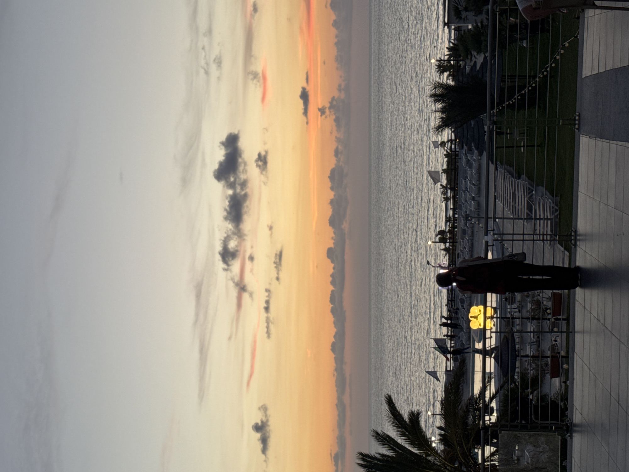 The image captures a twilight scene at a coastal resort, likely in Isola delle Femmine, Italy. The sky displays a gradient of soft oranges, yellows, and pale blues as the sun sets over the calm, grey sea. In the foreground, a person stands on a tiled balcony, silhouetted against the fading light, holding a phone up as if taking a picture. They are facing the sea, with their back to the viewer.
Below the balcony, a resort area is visible, with rows of empty lounge chairs and thatched umbrellas suggesting a beachside setting. Several flags, some white and some bearing emblems, are planted along the waterfront. Palm trees and other greenery are scattered throughout the resort. In the middle ground, there are several yellow, illuminated decorative objects, possibly lamps or art installations, near the water's edge. The overall atmosphere is serene and tranquil, marking the end of the day.