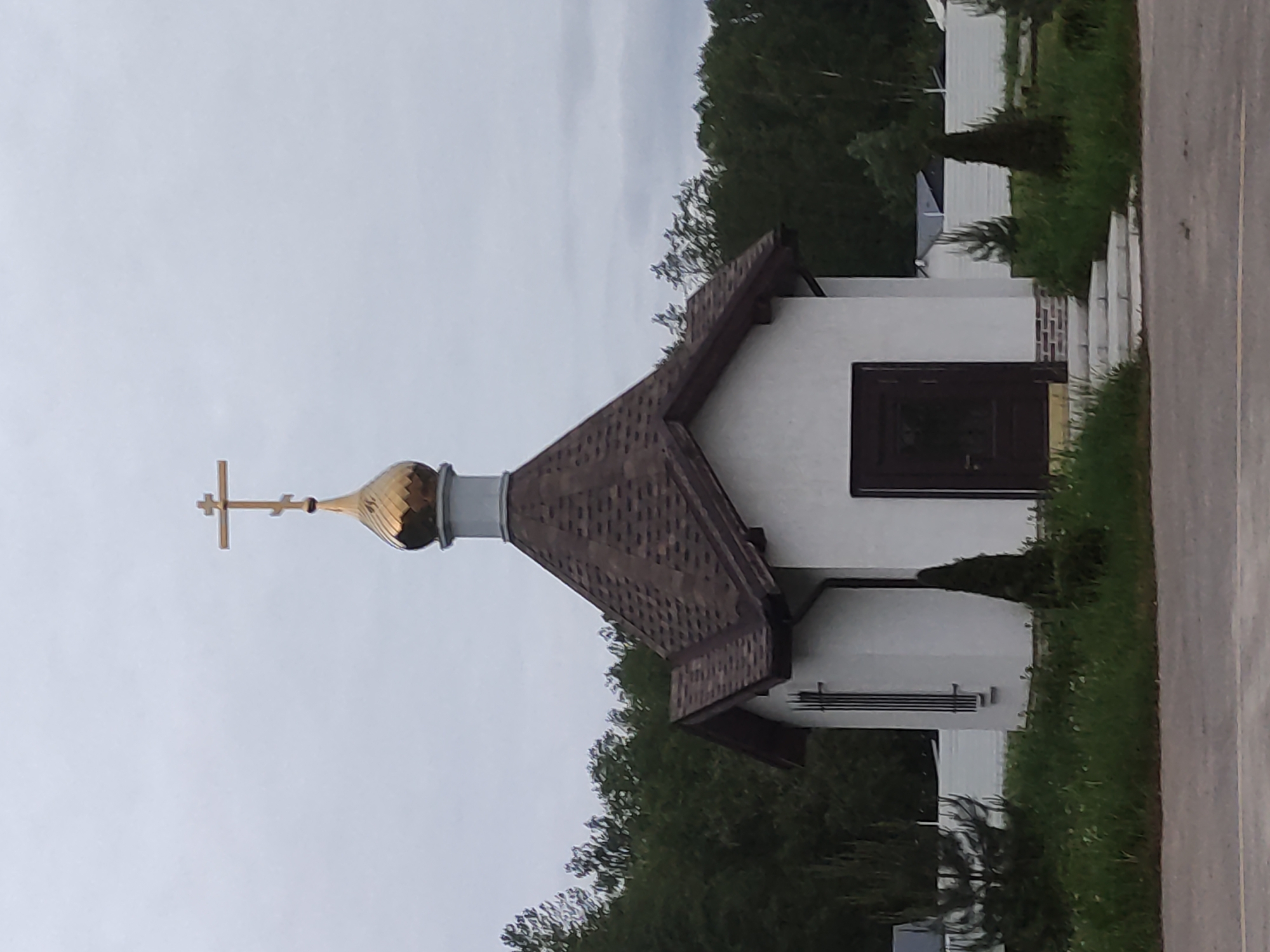 A building, possibly a small church or chapel, is depicted in Shevelkino, Russia. The building has a white exterior with a brown roof. A golden dome and cross adorn the structure. The surrounding environment includes greenery, suggesting a park-like or landscaped area. The sky overhead is overcast. A road is visible on the right. No people are visible in the frame.