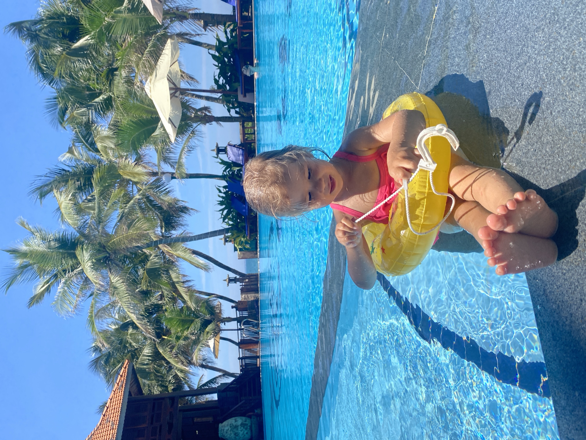 A young girl with blonde hair, wearing a pink swimsuit and a yellow inflatable ring, sits on the edge of a bright blue swimming pool. She is holding onto the rope of the inflatable ring, which is around her waist. Her bare feet are visible, with water droplets on them. The pool is surrounded by a dark gray tiled edge. In the background, palm trees sway against a clear blue sky. A glimpse of a building with a terracotta roof can be seen among the trees. The scene appears to be set during the daytime, under sunny weather, in a tropical location like Phu Thuy Ward, Vietnam. The girl's facial expression suggests happiness and engagement with her activity in the pool. There is no visible text in the image.
