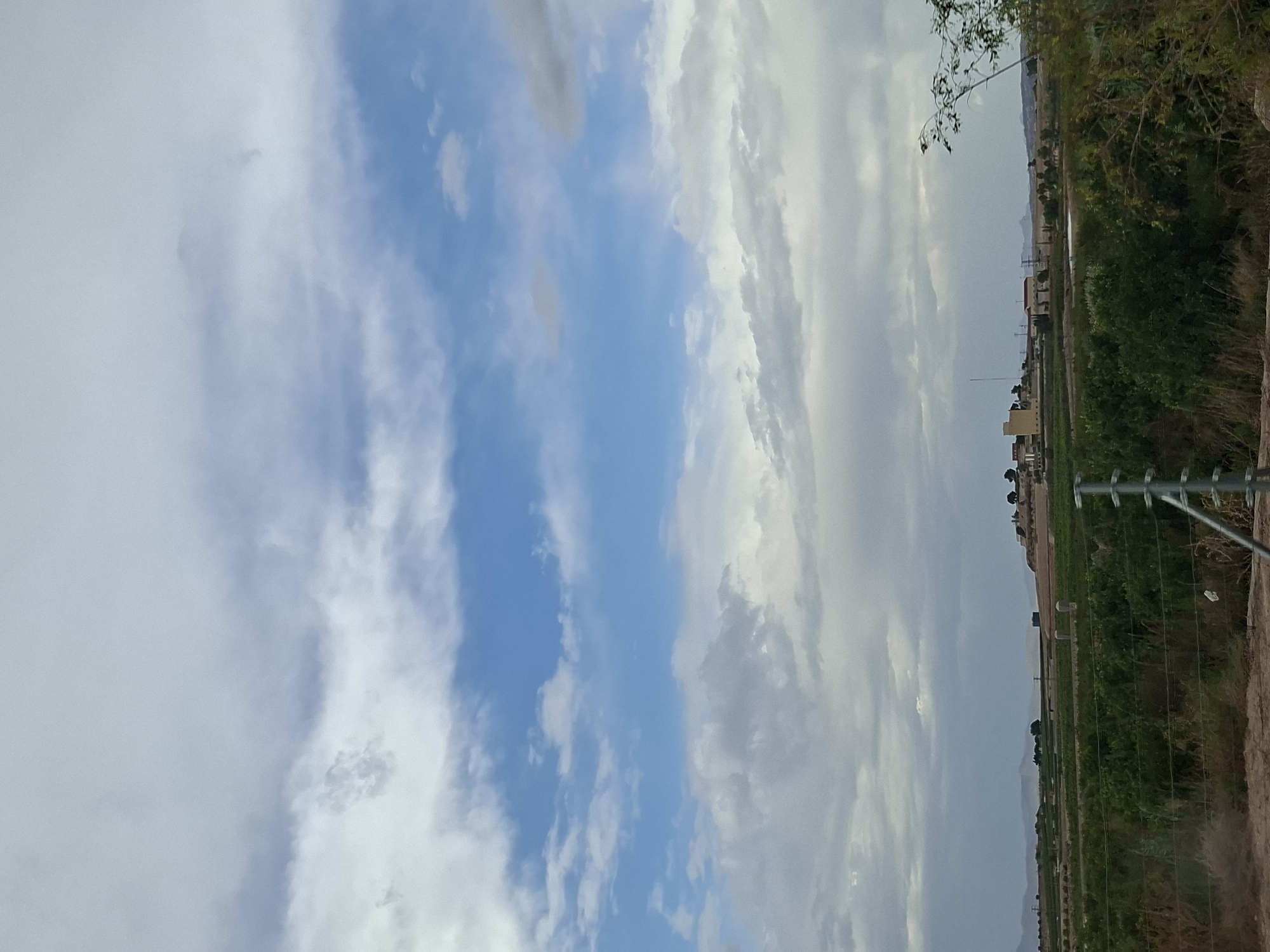 The image depicts a rural landscape under a cloudy sky. In the foreground, a metal fence runs horizontally across the frame, with dry brush and sparse vegetation around it. Beyond the fence, agricultural fields stretch into the distance, filled with rows of green crops. In the mid-ground, a cluster of buildings, including a prominent yellow tower, sits on a slight rise, surrounded by trees. Further in the background, a range of hazy mountains forms the horizon. The sky is a mix of blue patches and heavy gray clouds, suggesting an overcast or potentially stormy day. The scene appears to be in a semi-arid region.