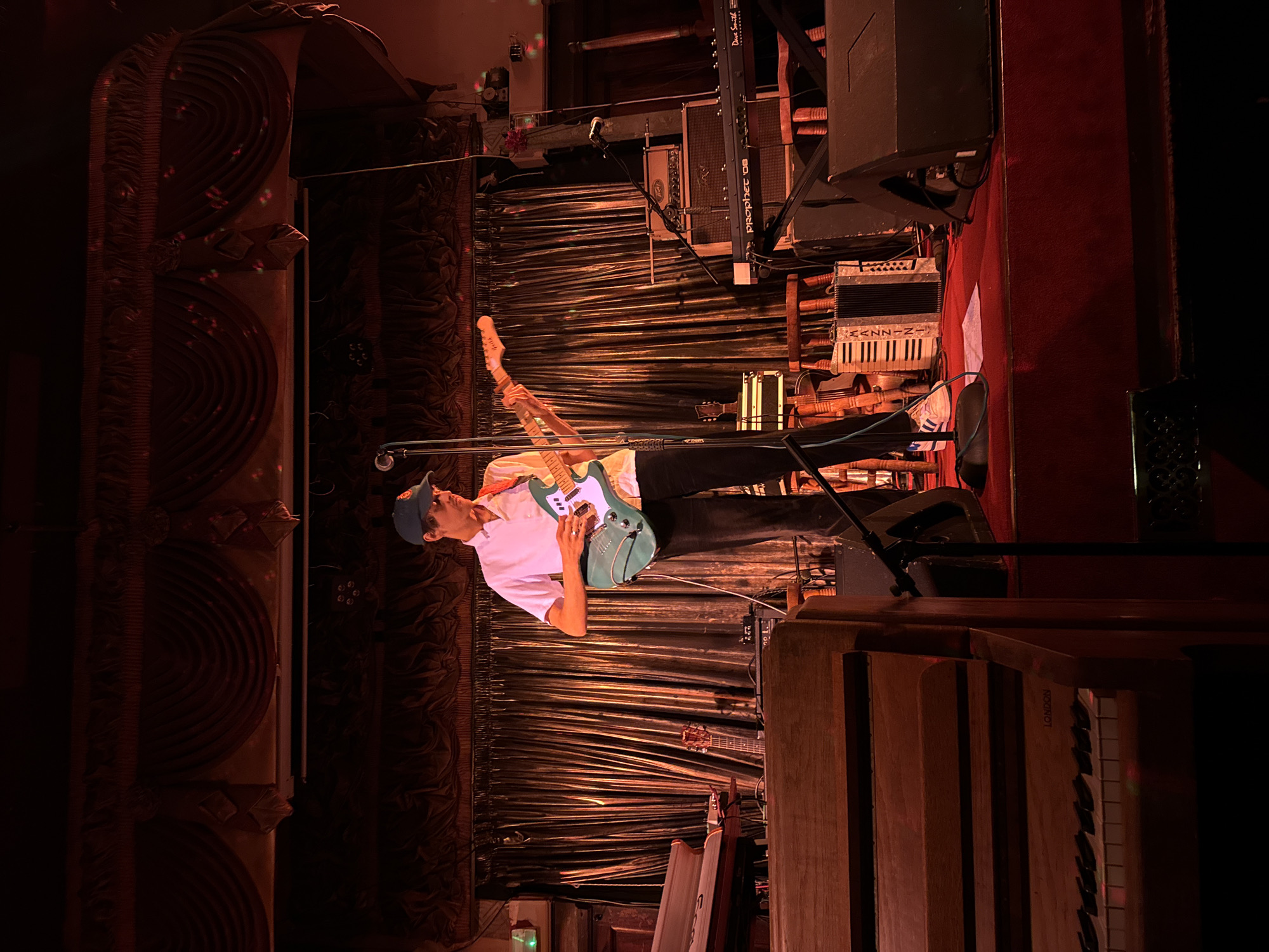 This rotated image depicts a live musical performance on a stage, likely at a venue in London, United Kingdom, given the text visible on an instrument. The scene is dimly lit with warm red and orange stage lighting, creating a dramatic atmosphere.
In the foreground, a male musician is captured mid-performance, standing and playing a light teal-colored electric guitar. He is dressed in a light-colored short-sleeved shirt, dark trousers, and a blue baseball cap, and is singing into a microphone on a stand in front of him. Behind the musician, the stage backdrop consists of dark, pleated fabric or curtains.
The stage is equipped with several instruments and audio gear. To the musician's right, an accordion with a keyboard and the partially visible brand "KASIN-N" or "KASSIN" rests on a stand. Above it, a synthesizer labeled "Prophet '08" by "Dave Smith Instruments" is mounted. Another guitar is visible on a stand further back. On the far right foreground, a large wooden instrument with a visible keyboard, possibly an organ, prominently features the word "LONDON" on its side. On the far left, a tall, decorative wooden structure with two large circular patterns suggests ornate venue architecture or specialized speaker enclosures. Various cables, stands, and speaker cabinets are also visible, contributing to the live music setting. The floor on the right side of the stage appears to be covered with red carpeting. The ambiance suggests an evening concert or musical event.