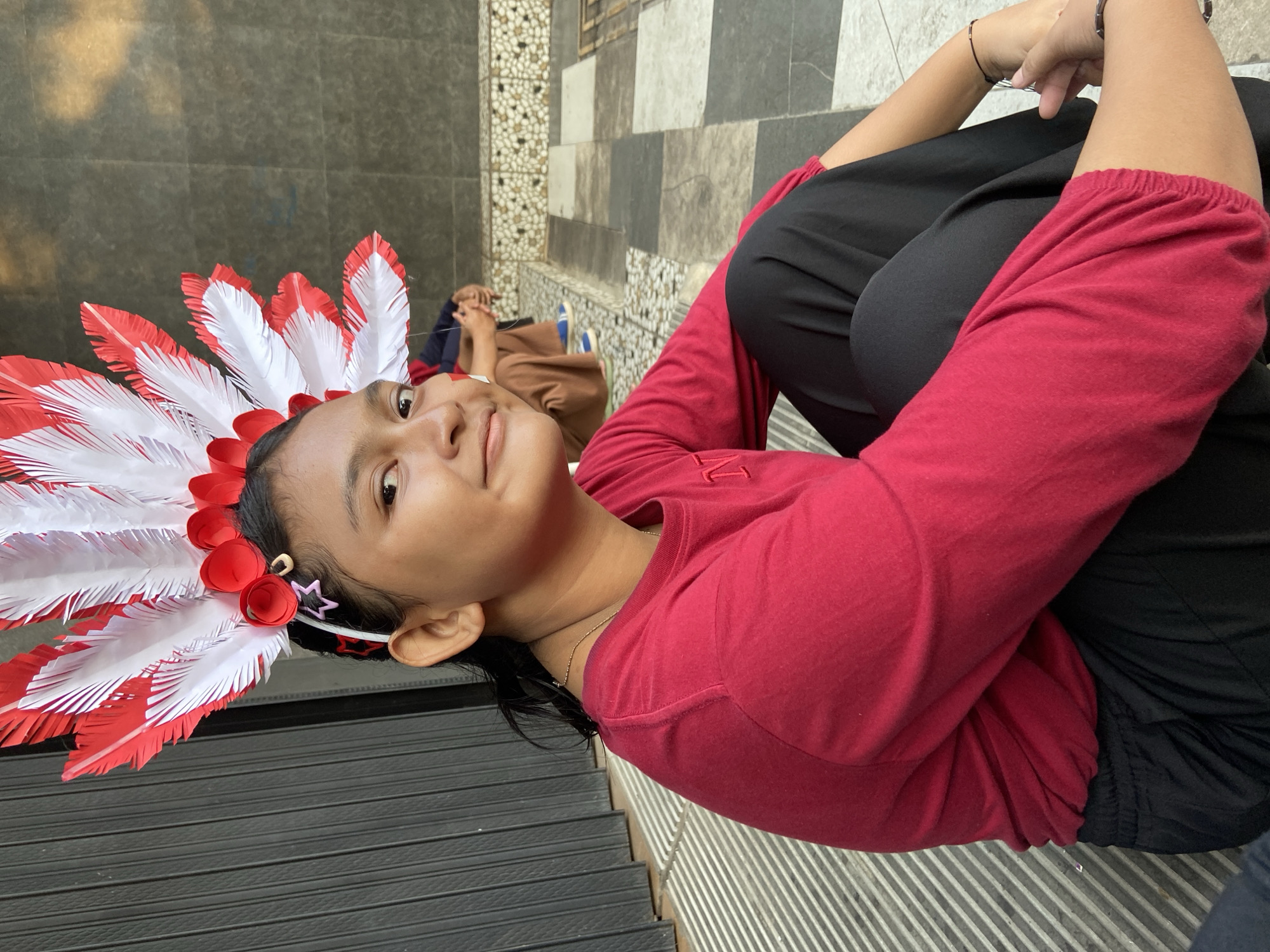 A young girl with dark hair pulled back and a headdress adorned with red and white feathers sits on steps outside a building. She is wearing a long-sleeved red shirt and black pants. Her left arm is resting on her lap, while her right hand is near her chin. The background shows a dark grey wall and a patterned tile border. In the distance, another person is seated on the steps. The scene appears to be in an outdoor urban setting, possibly in Kecamatan Pati, Indonesia, given the architectural elements and the presence of a gate on the left.