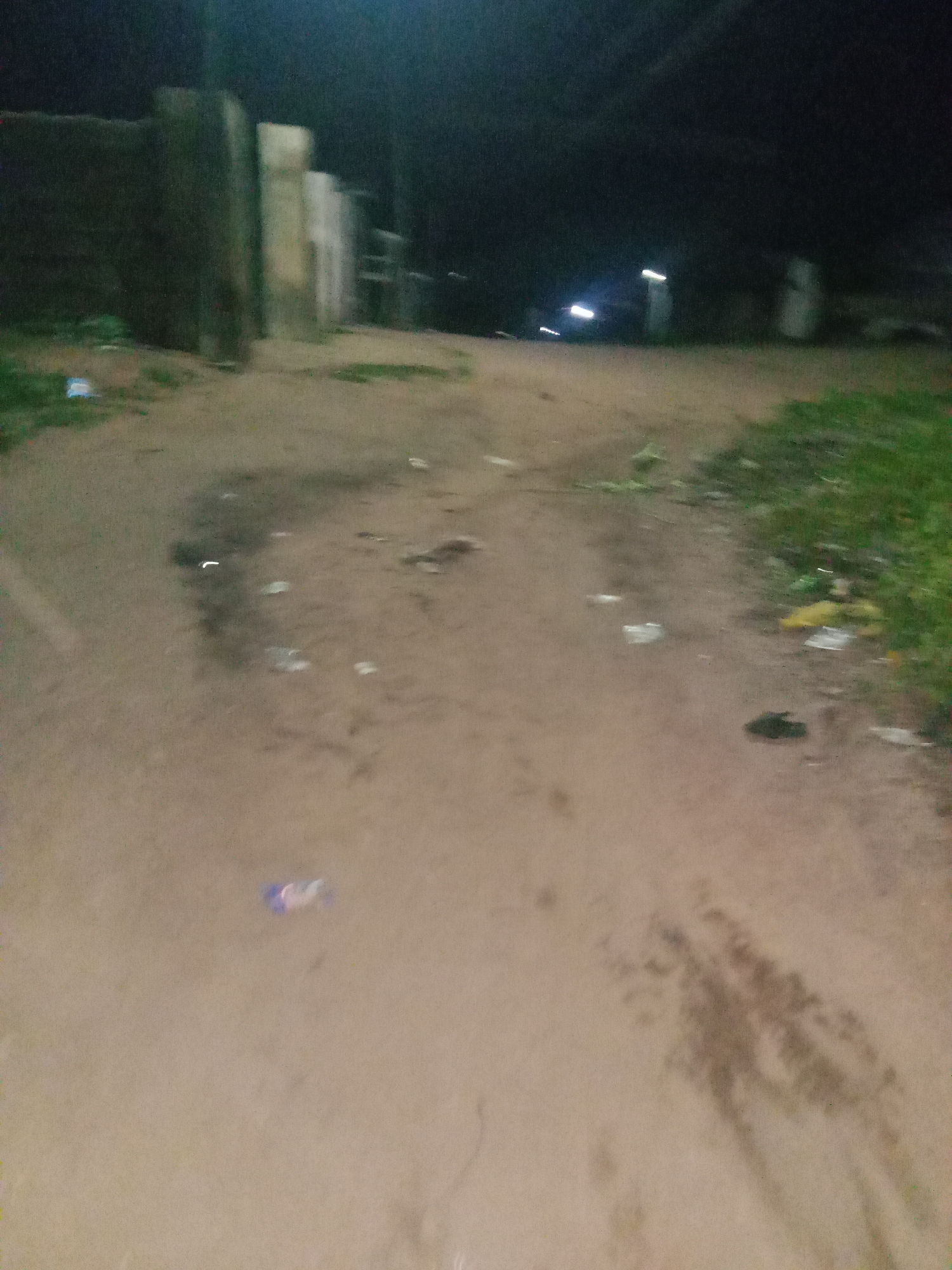 The image depicts a dimly lit, outdoor scene, likely at night, with an unpaved, sandy ground. The ground is littered with various pieces of debris, including plastic wrappers and what appear to be discarded food items. In the background, there are some structures, possibly walls or buildings, and faint lights from what could be vehicles or streetlights. The overall impression is of a somewhat neglected or unkempt area. The environment suggests a street or pathway in a residential or commercial area. There are no clearly identifiable individuals or specific actions taking place.
