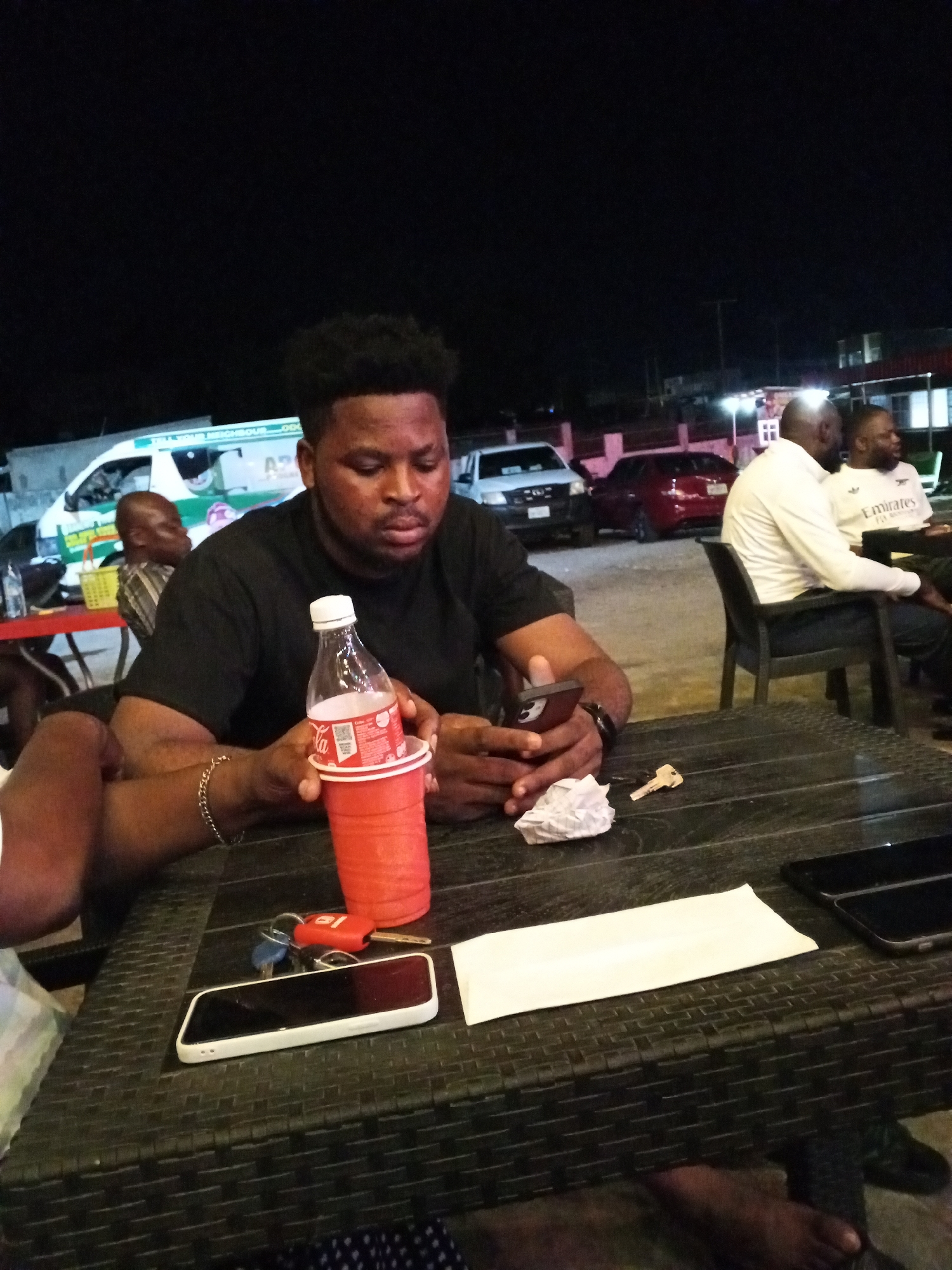 The image depicts an outdoor scene at night, possibly in Lokoja, Nigeria, although this cannot be definitively confirmed. A man is seated at a dark-colored wicker table, looking at his mobile phone. He has dark hair, a dark complexion, and is wearing a black t-shirt. A bottle of Coca-Cola is placed in a red plastic cup on the table. Also on the table are keys, a crumpled white tissue, a white smartphone, a folded white paper item, and a black smartphone. In the background, other individuals are seated at a separate table. A white ambulance is visible in the background, along with several cars parked in a lot. The lighting is artificial and appears to be coming from overhead lights.