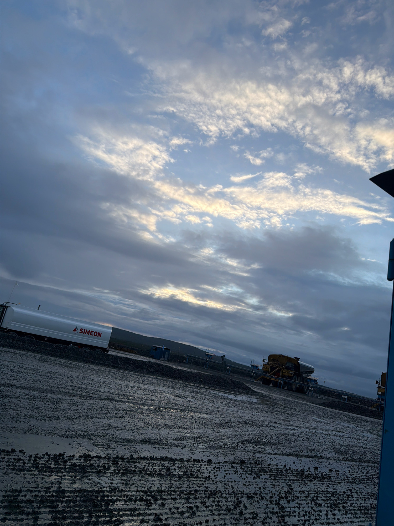The image depicts an outdoor scene, likely an industrial area, under a cloudy sky. In the foreground, a wet, gravelly surface is visible, with tire tracks indicating recent vehicle activity. To the left, a white tanker truck with "SIMEON" written in red lettering is parked. Further in the background, a large yellow mining truck is seen, along with other industrial structures and equipment. The overall impression is that of a worksite, possibly a mine or a transportation hub, in the Gällivare region of Sweden, given the context provided. The lighting suggests it may be dawn or dusk. There are no people visible in the image.