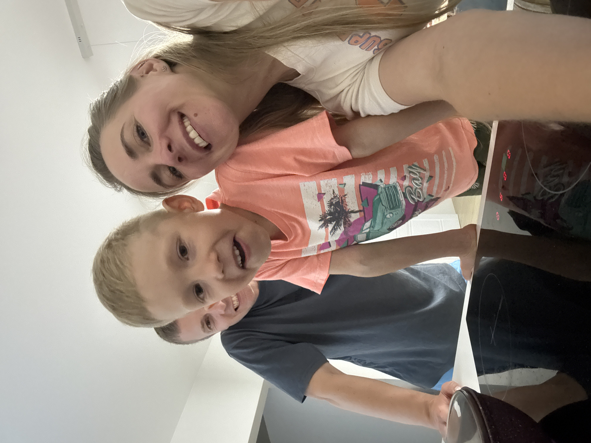 The image captures a low-angle selfie of three smiling individuals in a modern indoor setting, likely a kitchen. The perspective indicates the camera is held by the woman on the right.
On the left, an adult male with short, light-brown hair and a light stubble is partially visible from the chest up. He wears a dark grey short-sleeved t-shirt and is leaning forward with his forearms resting on a white countertop. He smiles broadly, revealing his teeth.
In the center, a young boy, approximately 4-7 years old, with short blonde hair, stands slightly in front of the man. He wears an orange short-sleeved t-shirt adorned with a multi-colored graphic print depicting palm trees and a classic-style vehicle at a beach. The boy smiles with an open mouth, looking directly at the camera. His left forearm is partially visible, resting on the white countertop.
On the right, an adult female with long, light-blonde hair, partly pulled back, is positioned closest to the camera. She wears a light-colored (off-white) short-sleeved t-shirt with an indistinct partial orange print on the chest. She smiles widely, showing her teeth, and has a small dark mole on her right cheek. Her visible ear features multiple small earrings, including a helix piercing. Her right arm is angled towards the bottom right, suggesting she is holding the camera for the selfie.
In the immediate foreground, a dark, reflective induction stovetop is visible, featuring circular burner outlines and illuminated red digital displays. A dark, possibly maroon, pot or bowl is partially visible on the far left of the white countertop next to the stovetop.
The background consists of plain, light-colored walls and a white ceiling. A modern, linear light fixture is mounted on the ceiling in the upper right. A darker vertical element, possibly a door frame or cabinet edge, is present on the far left.
Based on the visual evidence within the image, it is not possible to confirm that the location is the city of Chaikovsky, Russia. The environment depicts a generic modern residential interior.