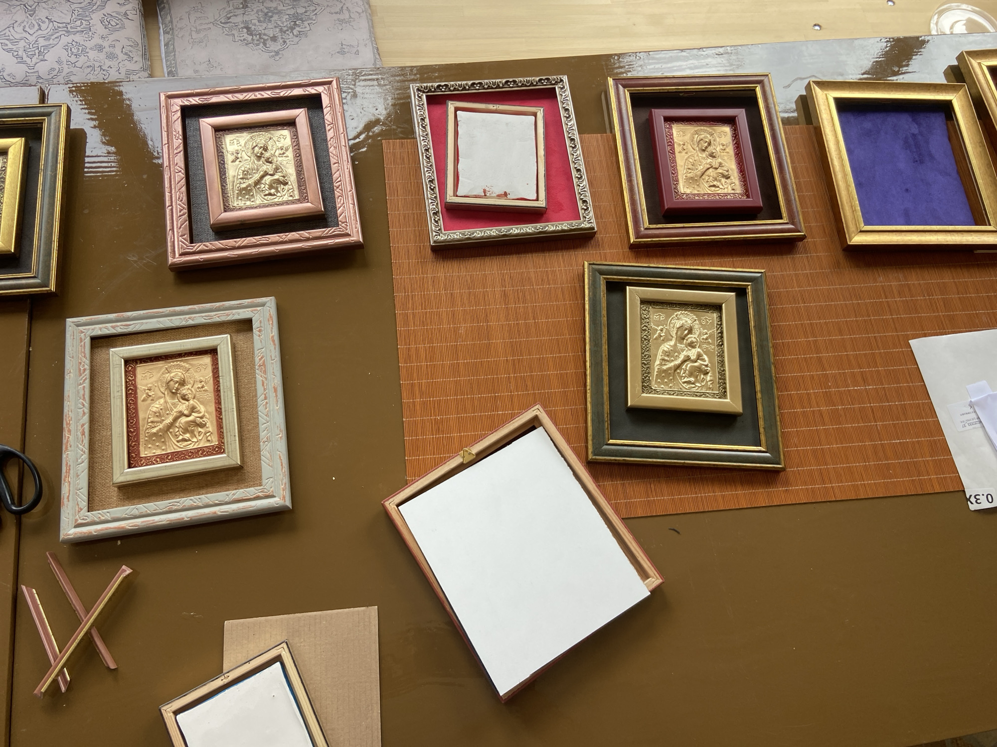 The image shows a collection of framed religious icons and empty frames on a brown surface, part of which is covered with a bamboo-like mat. The icons are of Byzantine or Eastern Orthodox style, with golden figures on a dark background, framed with different materials and colors, including gold, brown, and grey. Some frames are empty, showing either white or colored backing. There are scissors and other materials on the brown surface. The location in Donji Katun, Montenegro is unknown.