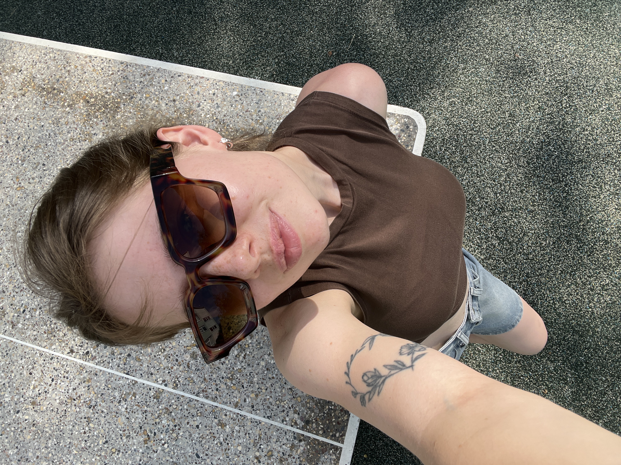 An overhead medium shot captures a young Caucasian woman wearing large, brown-patterned sunglasses, a brown tank top, and denim shorts. She is lying on her back on a speckled grey surface with a white line marking a boundary. To her right, the surface transitions to a dark grey, textured ground. The woman has light brown hair pulled back and is wearing small silver hoop earrings. A tattoo of a floral wreath is visible on her left forearm. The sun is shining, casting shadows and highlighting the texture of the surfaces. The scene appears to be an outdoor recreational area, possibly a park or sports court. No specific location in Chaikovsky, Russia is identifiable from the image itself.