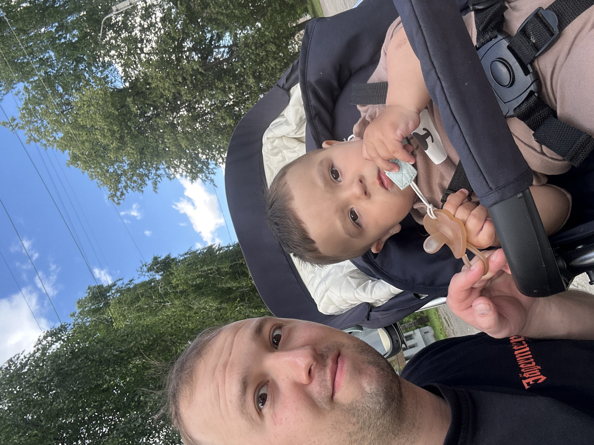 The media file shows a selfie of a man and a baby in a stroller, taken outdoors in Chaikovsky, Russia. The man, with short brown hair and a beard, is in the foreground, looking at the camera. The baby, positioned in a stroller behind the man, has dark hair and is looking to the side, holding a pacifier. The baby is wearing a light brown onesie. The stroller has a dark blue fabric and a white padded insert.
The background consists of trees with green foliage, a bright blue sky with some white clouds, and overhead power lines. The lighting suggests it is daytime, likely mid-morning or afternoon, with bright sunlight. The overall scene depicts a casual outdoor moment, possibly a walk in a park or along a street. The expressions of both individuals appear neutral. Visible text on the man's black t-shirt reads "Jägermeister" in a stylized font.