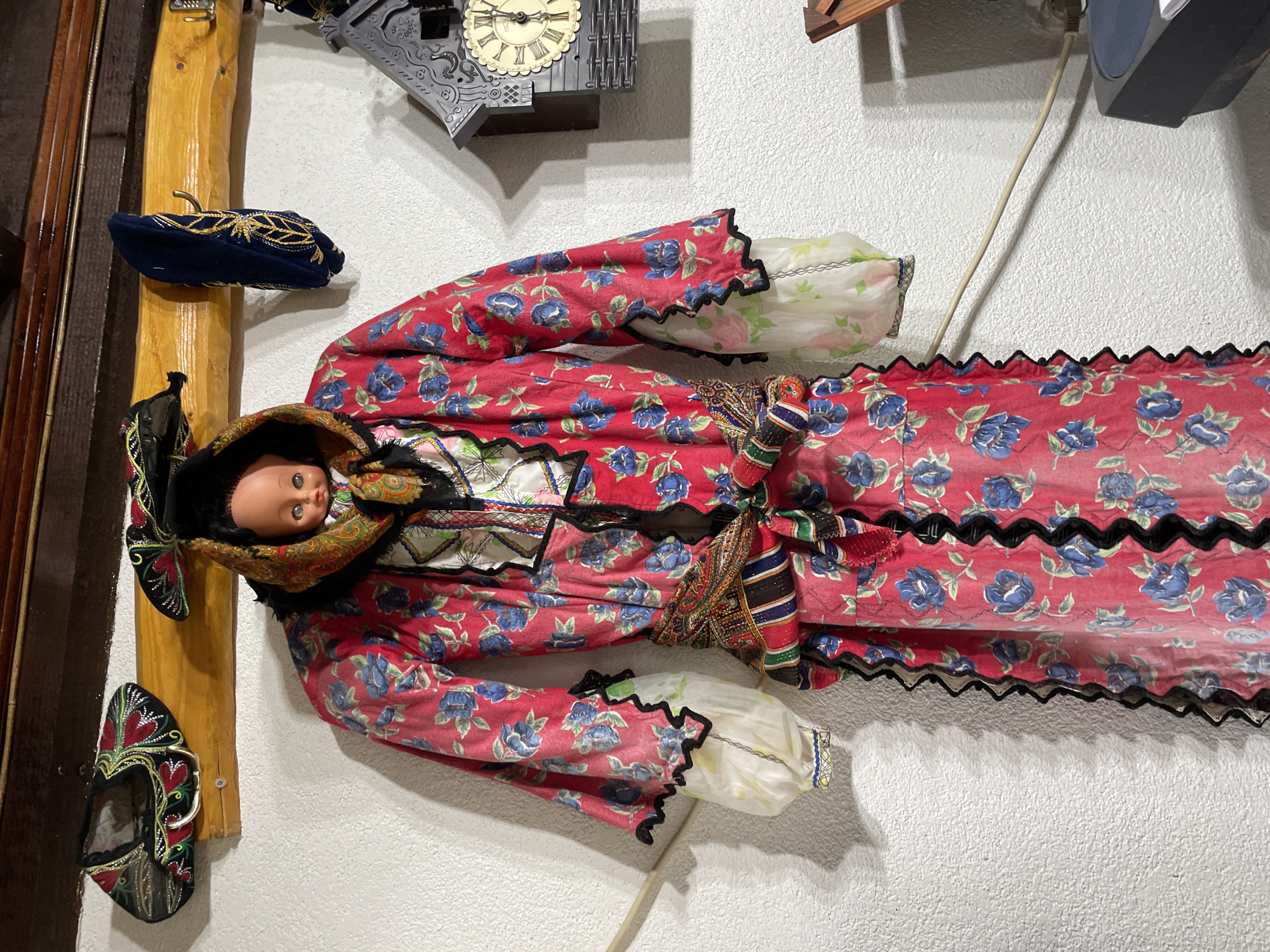 A doll dressed in traditional Bulgarian clothing is mounted on a wall alongside various other decorative objects. The doll's dress features a red floral pattern with blue flowers, accented by black zig-zag trim. A multi-colored sash is wrapped around the waist. The doll has a fair complexion, and its head is adorned with a traditional hat and shawl. Next to the doll are a wooden frame with a blue hat and a shoe also in Bulgarian traditional clothing. Above the doll is a cuckoo clock. The overall setting suggests an interior space, possibly a museum or a home displaying cultural artifacts, likely in Zlatograd, Bulgaria.