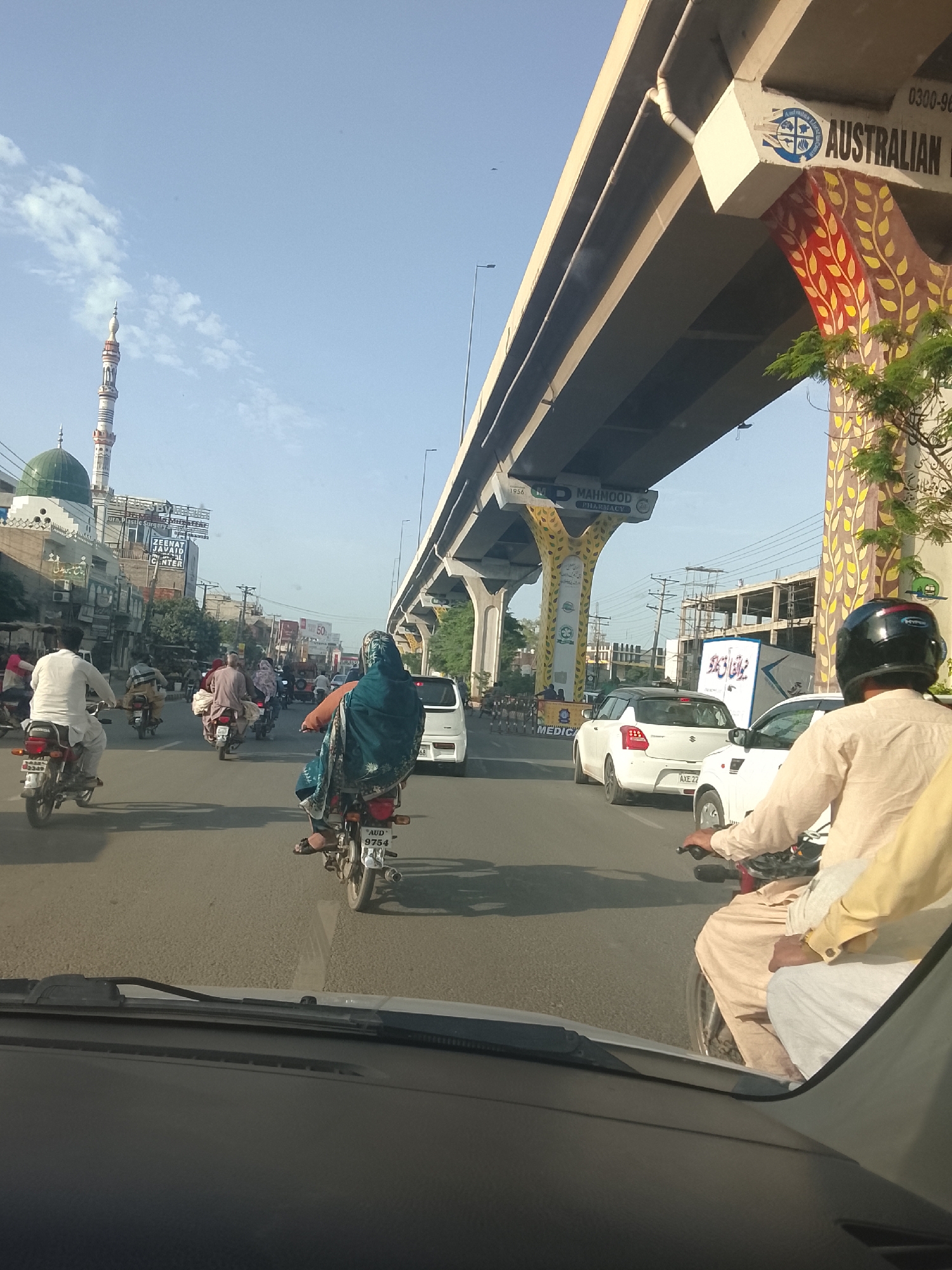 The image depicts a busy street scene in an urban area, possibly in Pakistan. Several people are riding motorcycles, some wearing traditional clothing. There is a large overhead bridge structure with the words "AUSTRALIAN" visible. A mosque with a green dome and a tall minaret can be seen in the background. Numerous vehicles are present, including cars and motorcycles, navigating the road. The view is taken from inside a car, looking out through the windshield. Signboards and shopfronts are visible in the background, including one for "ZEENAT JAVAID MEDICAL CENTER" and "MAHMOOD PHARMACY."