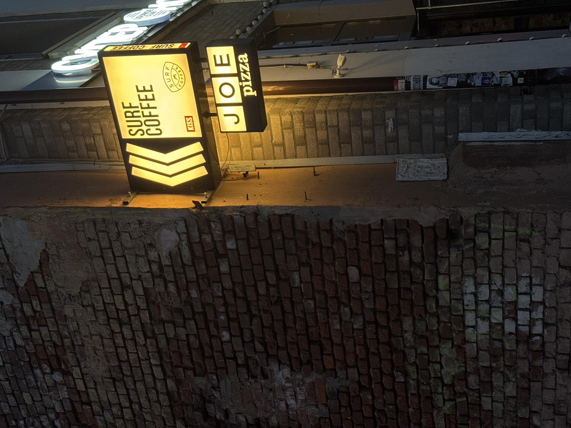 The image shows a storefront at night in Moscow, Russia. Two illuminated signs are visible: one reads "SURF COFFEE" with a logo and "KISS" underneath, and the other reads "JOE pizza." The signs are attached to a brick wall, which appears to be part of an older building. To the left, a rough brick wall occupies much of the frame, showing signs of age and wear, with some sections appearing to have peeling plaster or paint. To the right, a lighter colored section of the building is visible, with a doorway and some posters or stickers on the wall next to it. The lighting suggests it is nighttime, and the signs are the primary light sources in the immediate vicinity. There are no people visible in the frame.