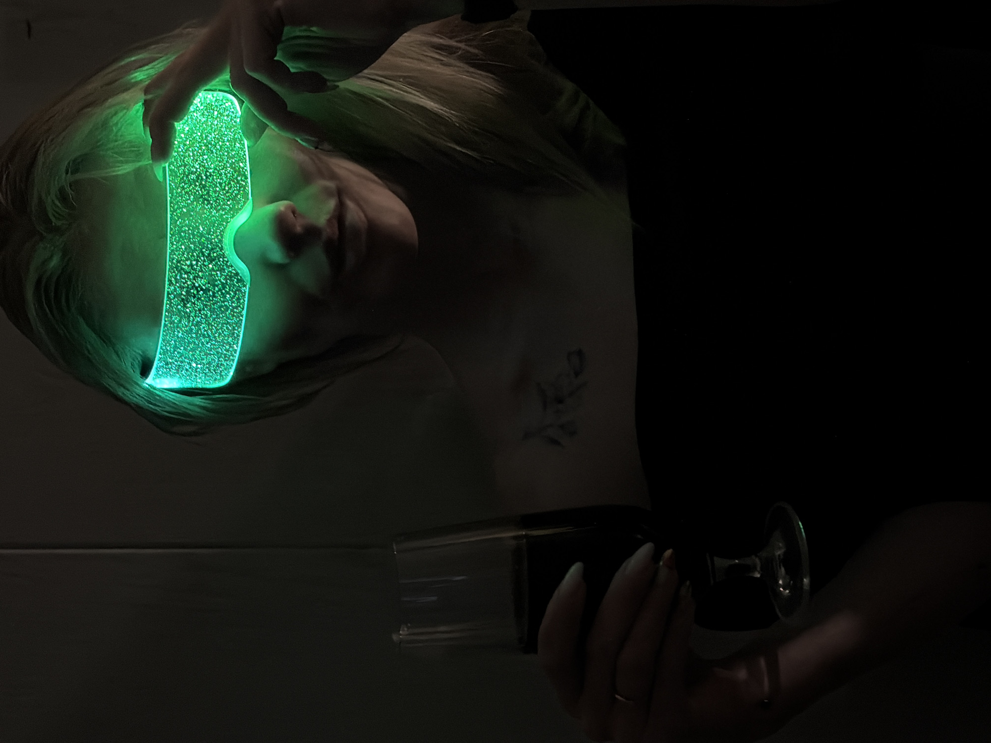 A woman with blonde hair is holding a glowing green visor over her eyes. She is also holding a black bottle with a clear liquid and a glass in her other hand. The scene is dark and dimly lit, with the green visor providing the primary light source on the woman's face. The woman has a tattoo on her chest. The location is unknown.