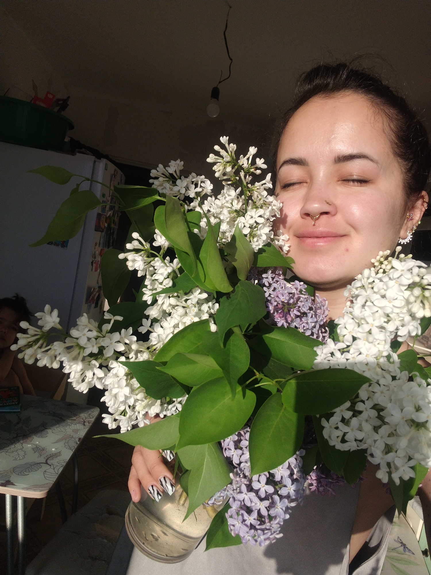 A woman is holding a bouquet of white and purple lilacs with green leaves. She is fair-skinned with dark hair pulled back, a nose ring, and earrings. Her eyes are closed and she has a slight smile. Her nails have a black and white zebra print. The bouquet is in a metallic vase. Behind her, there is a refrigerator, a table, and another person visible in the background. The setting appears to be indoors, possibly a kitchen.
