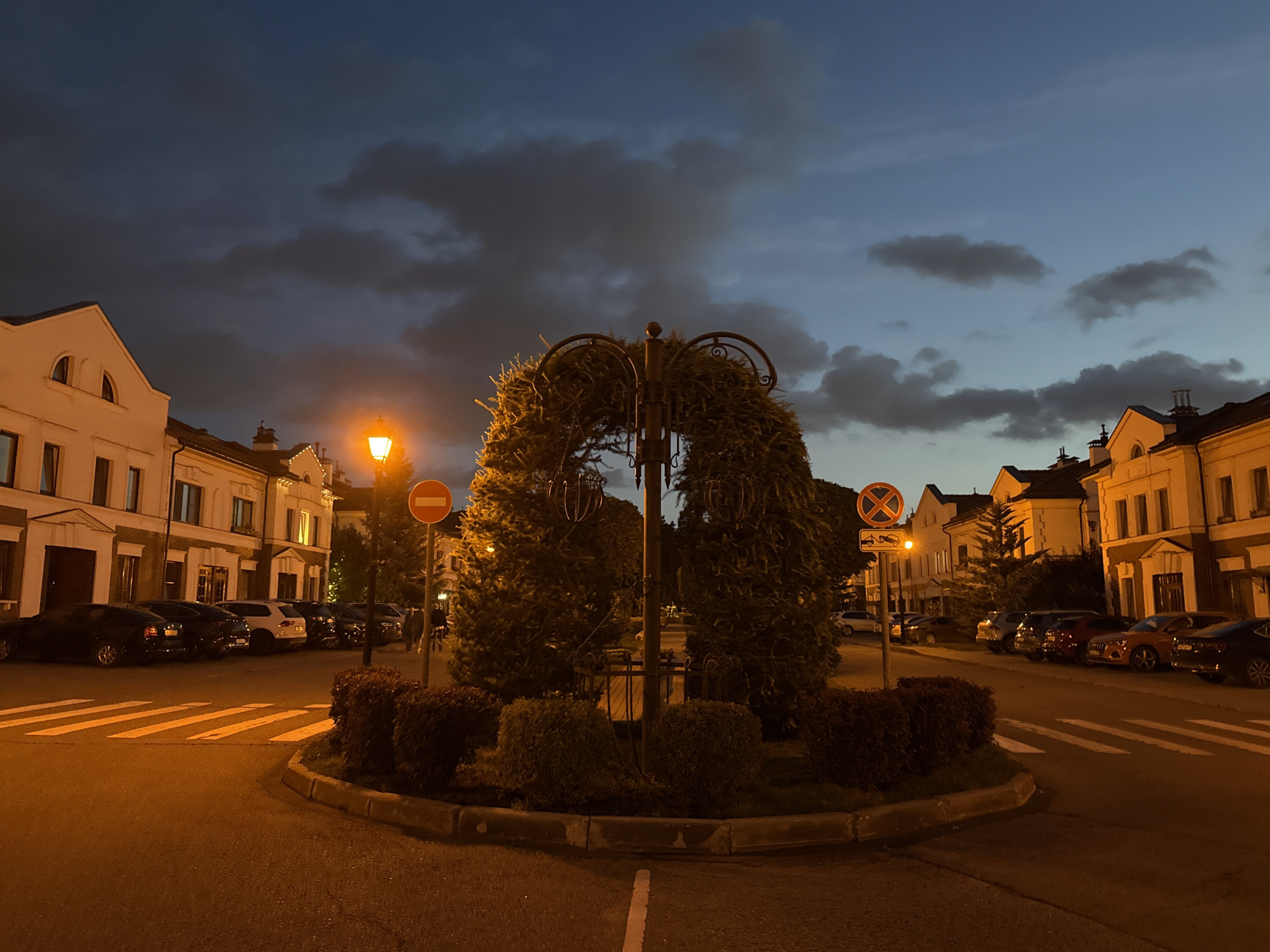 The image depicts a twilight scene in Lopatino, Russia, at a roundabout. In the center of the roundabout, a large, arched structure covered in foliage stands, reminiscent of a wreath, supported by a dark metal pole. Surrounding this central feature are neatly trimmed bushes. Two traffic signs are visible: one red and white prohibition sign, and another circular sign with a red border and a white background with black symbols.
The roundabout is paved with asphalt and features white pedestrian crossing lines. Several cars are parked along the side of the road on the left and right of the roundabout. The buildings lining the street are two-story structures with cream-colored facades and dark roofs, illuminated by warm, ambient light. The sky above is a deep blue transitioning to darker shades, with scattered clouds that are partially illuminated. A street lamp with a glowing orange light is visible on the left side of the image. The overall impression is one of a quiet, European-style residential area at dusk.