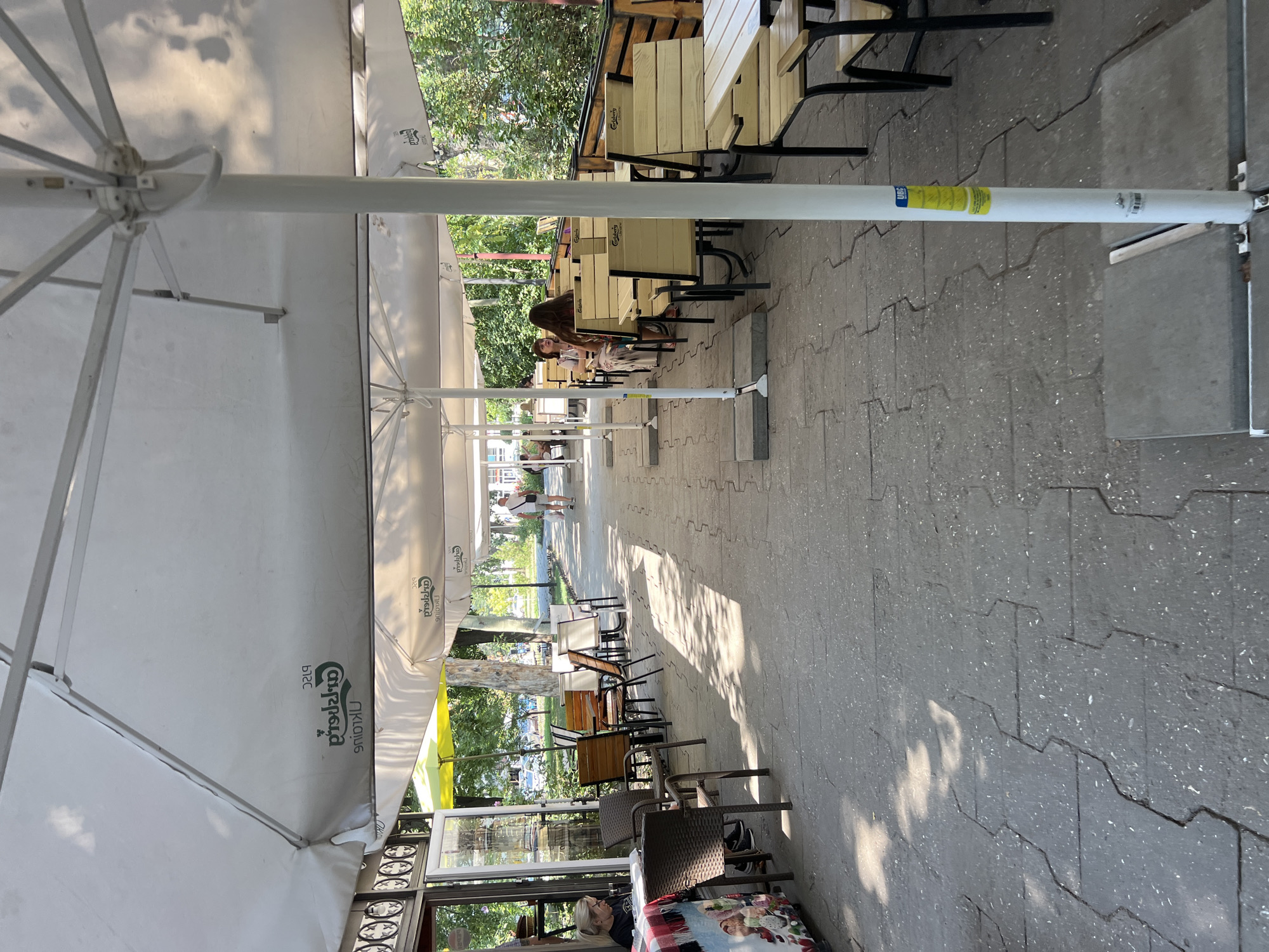 The image depicts an outdoor seating area of a cafe or restaurant on a sunny day in Odesa, Ukraine. Large white umbrellas provide shade over rows of tables and chairs arranged on a paved walkway. In the foreground, to the left, a woman with blonde hair is seated at a table near the entrance of a building, looking down at something on the table. To the right, the outdoor seating area extends further back, with several tables occupied by patrons. Further down the walkway, a man wearing a white t-shirt and shorts is walking away from the camera. The background shows trees and foliage, suggesting a location with greenery. The pavement is made of interlocking stone tiles, and there are shadows cast by the umbrellas and trees. The overall scene conveys a relaxed atmosphere of an outdoor dining establishment.