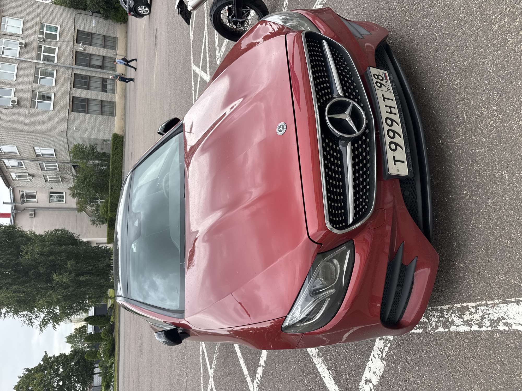 A red Mercedes-Benz E-Class coupe is parked in a lot with a building in the background. The car is facing forward, and its headlights are on. The license plate is visible and reads "T999HT 98 RUS". The car is parked in a parking spot marked with white lines. There are trees and a building with windows in the background. Two people are walking in the distance near the building. The sky is overcast.