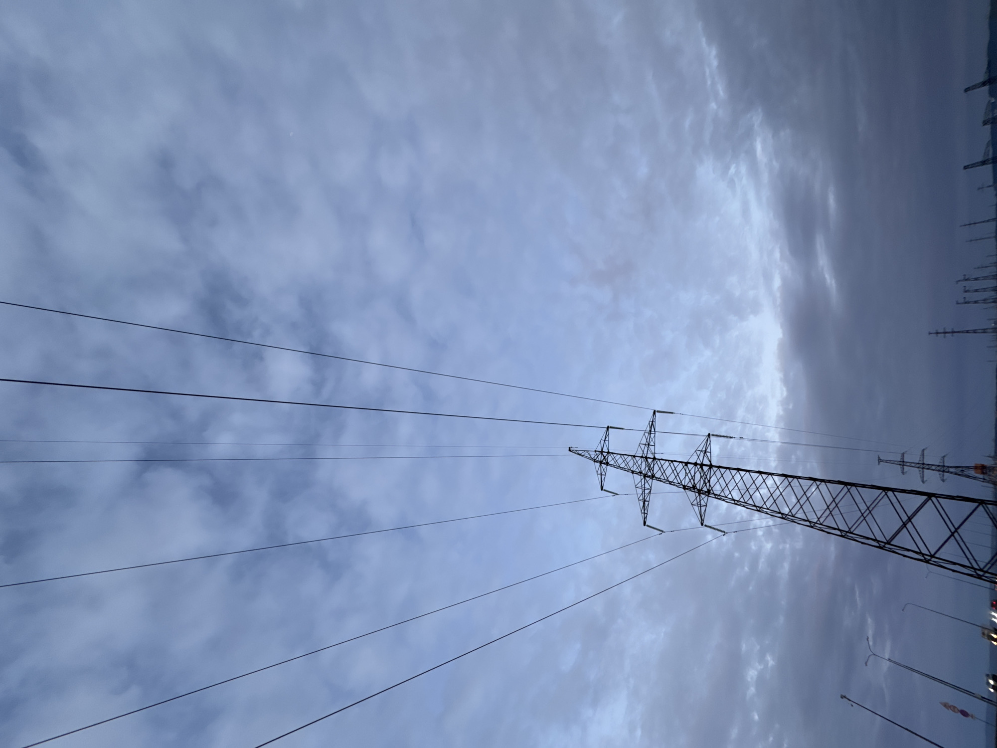 The image displays a large, dark metal lattice-work electricity transmission pylon positioned from the lower-left to the mid-left of the frame, extending upwards diagonally. Numerous power lines emanate from the pylon's cross-arms, stretching across a predominantly overcast sky. The sky is characterized by an extensive covering of blue-grey stratocumulus or altocumulus clouds. A faint, circular, pale celestial object, consistent with the moon, is visible in the upper-right quadrant. Along the distant horizon, in the lower right, several smaller, silhouetted electricity pylons are discernible, alongside indistinct industrial structures or cranes. On the far left, at ground level, artificial light sources resembling streetlights or vehicle headlights are visible, accompanied by a partial view of a red and white striped object. The lighting is subdued, indicating overcast conditions, possibly during twilight. This scene is located in the city of Chaikovsky, Russia.