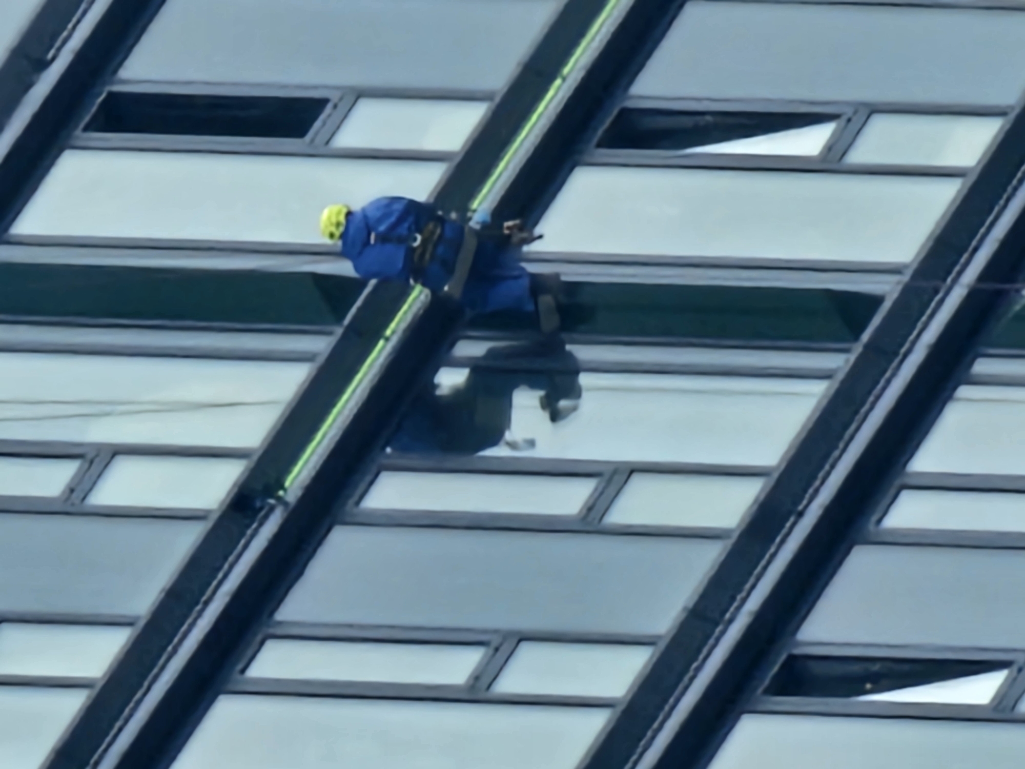A person in a blue uniform and yellow helmet is rappelling down the side of a modern building with a glass facade. The person is attached to ropes and appears to be performing maintenance or cleaning. The building has a sleek, contemporary design with dark framing around the large windows. The environment suggests an urban setting, likely a city, but no specific landmarks are visible to determine the exact location within Moscow, Russia. The lighting indicates it is daytime.